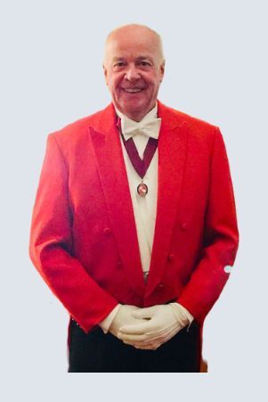 Man in a red jacket, white tie, gloves, and medal, smiling with hands clasped.