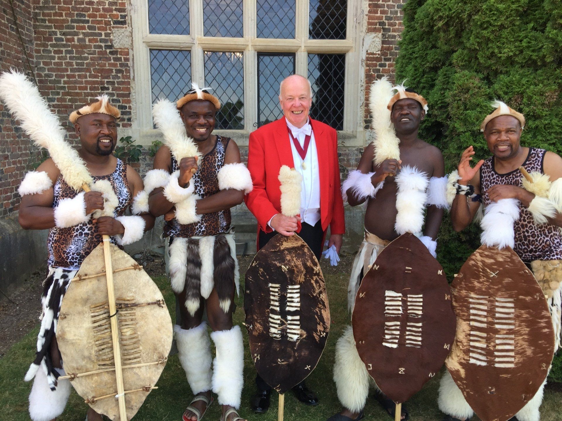 Man in red coat with four Zulu dancers holding shields and spears in front of a building.