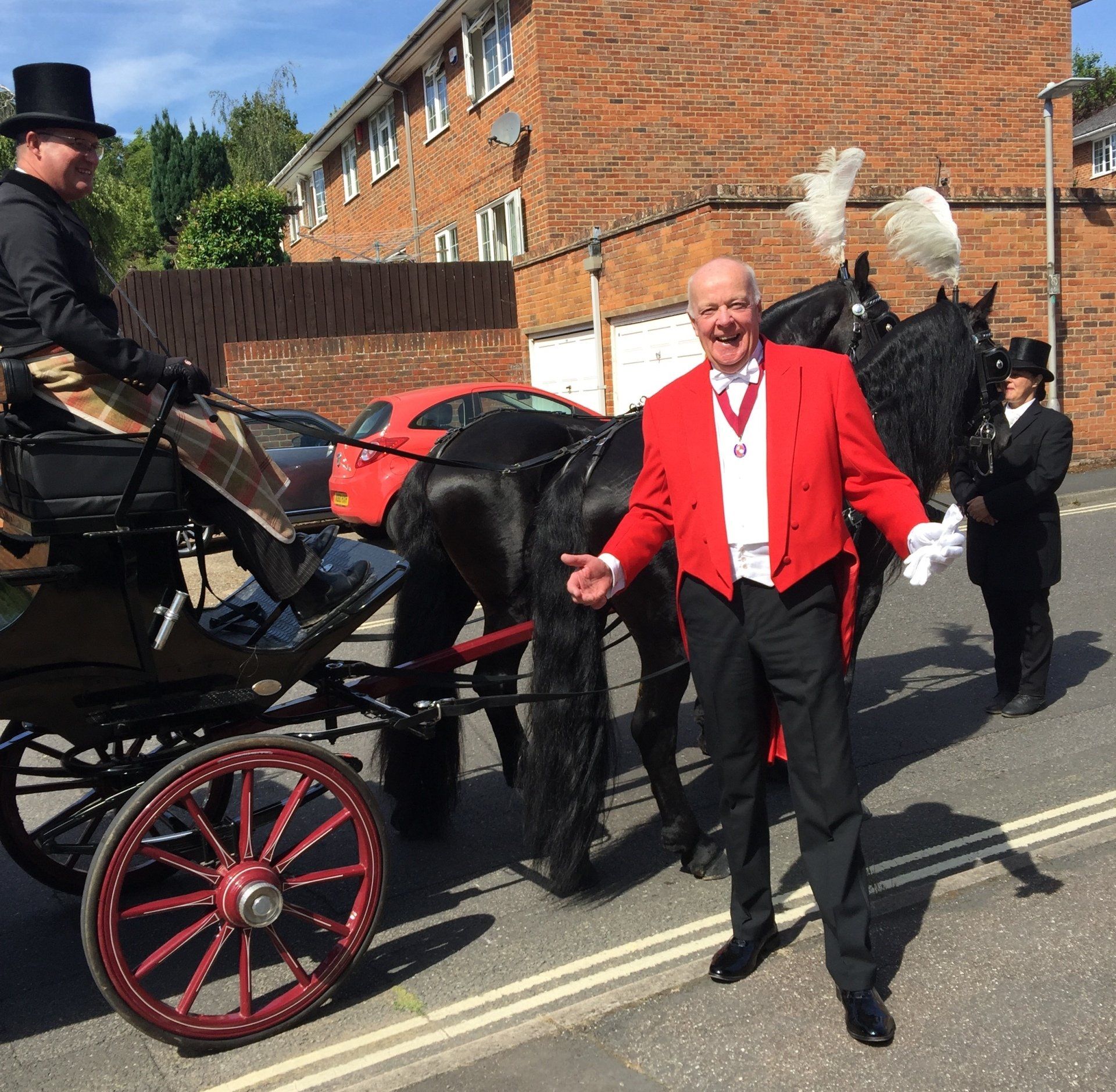 Man in red suit stands by horse-drawn carriage. Other men in formal attire. Sunny outdoor setting.