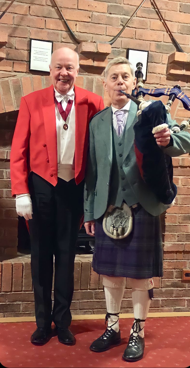 Two men posing in front of a brick wall. One in a red jacket, the other in a kilt playing bagpipes.