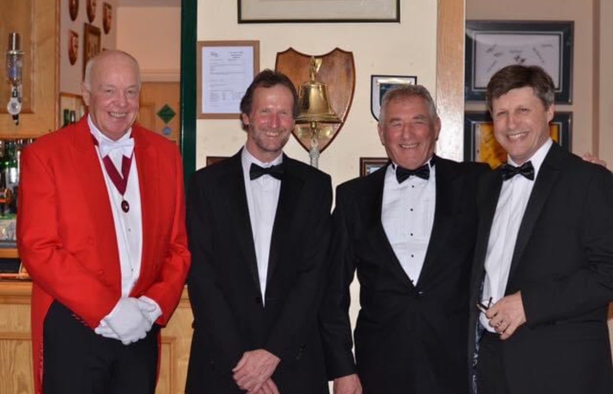 Four men in formal attire, one in red jacket, posing indoors, smiling.