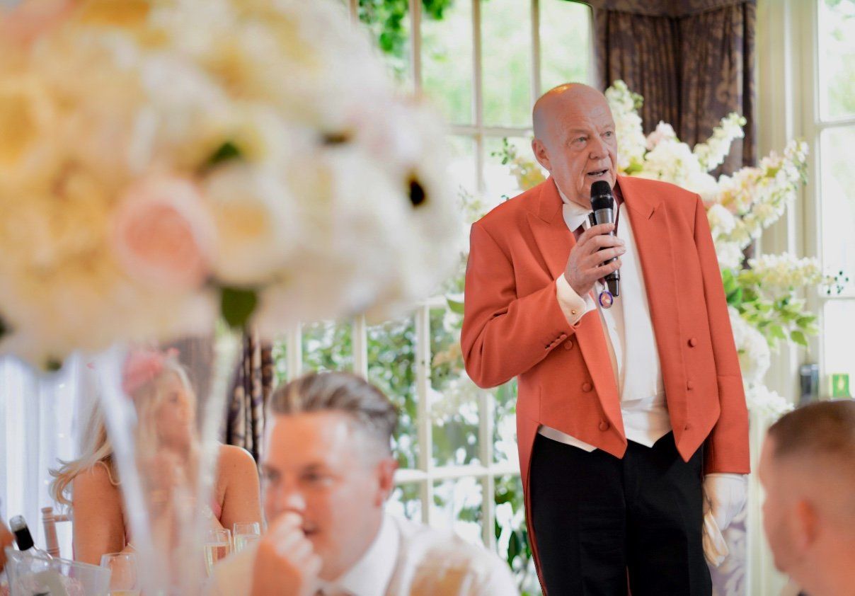 Man in orange jacket speaks into microphone at a wedding reception. Flowers and guests in the background.