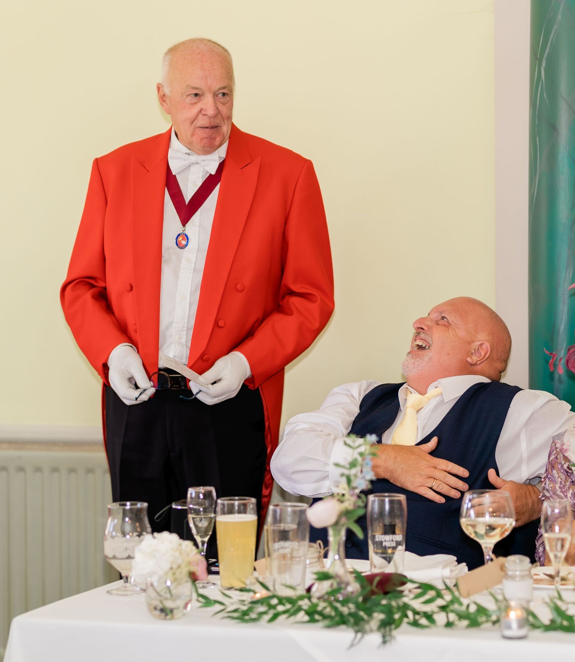 Man in red jacket stands near seated man laughing at a table with drinks and flowers.