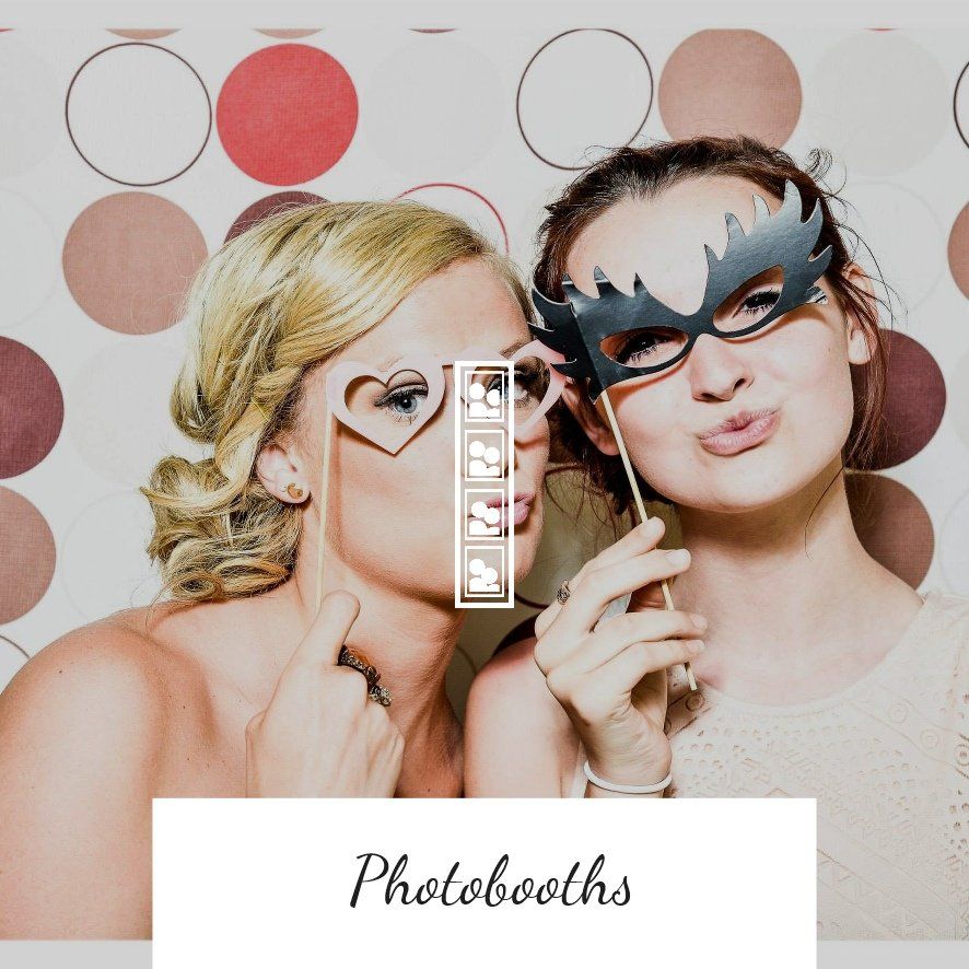 Two women pose with masks at a photobooth, making playful faces. They are in front of a backdrop with circles.