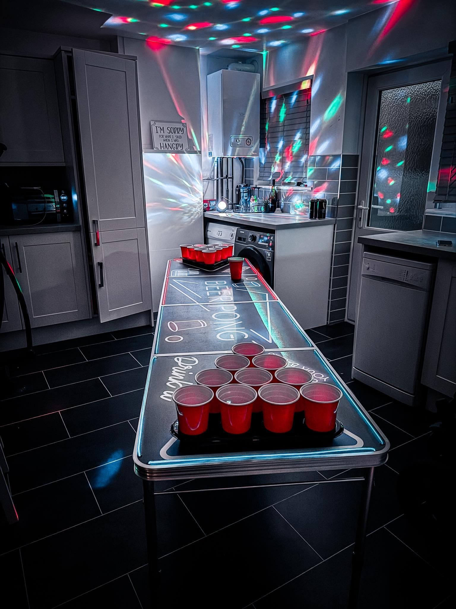 A kitchen with a beer pong table set up, with red cups arranged in a triangle, lit by disco lights.