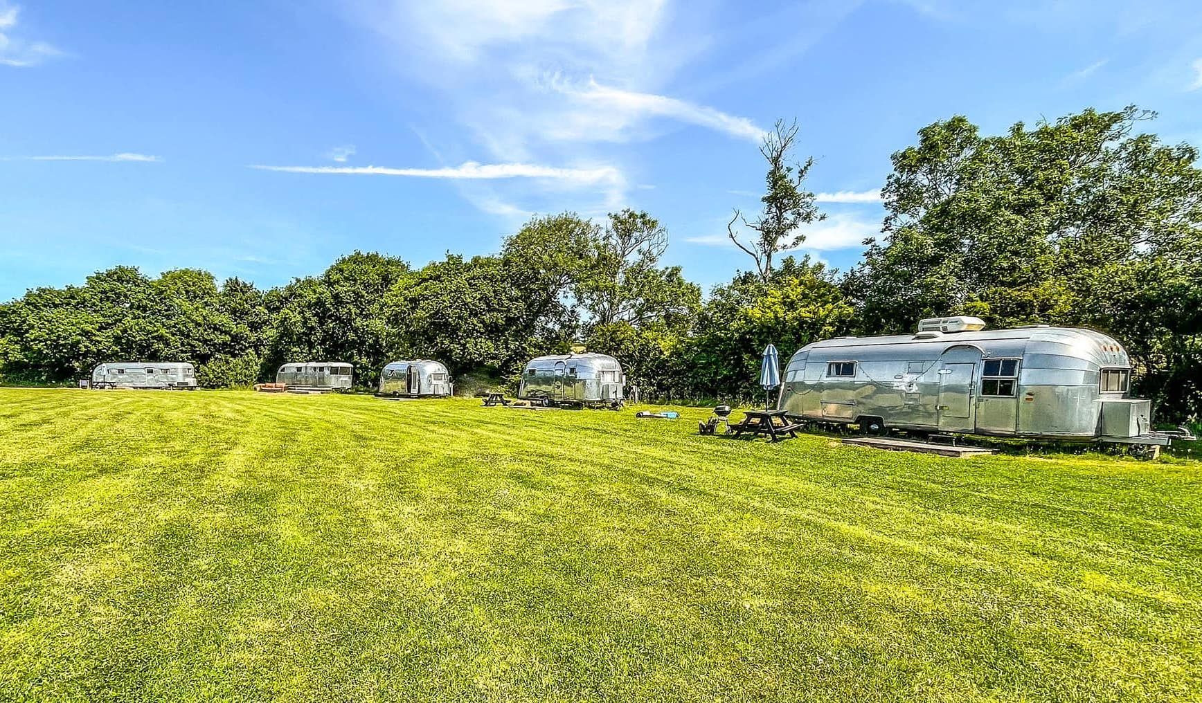 Silver travel trailers parked on green grass field, trees in the background, blue sky overhead.