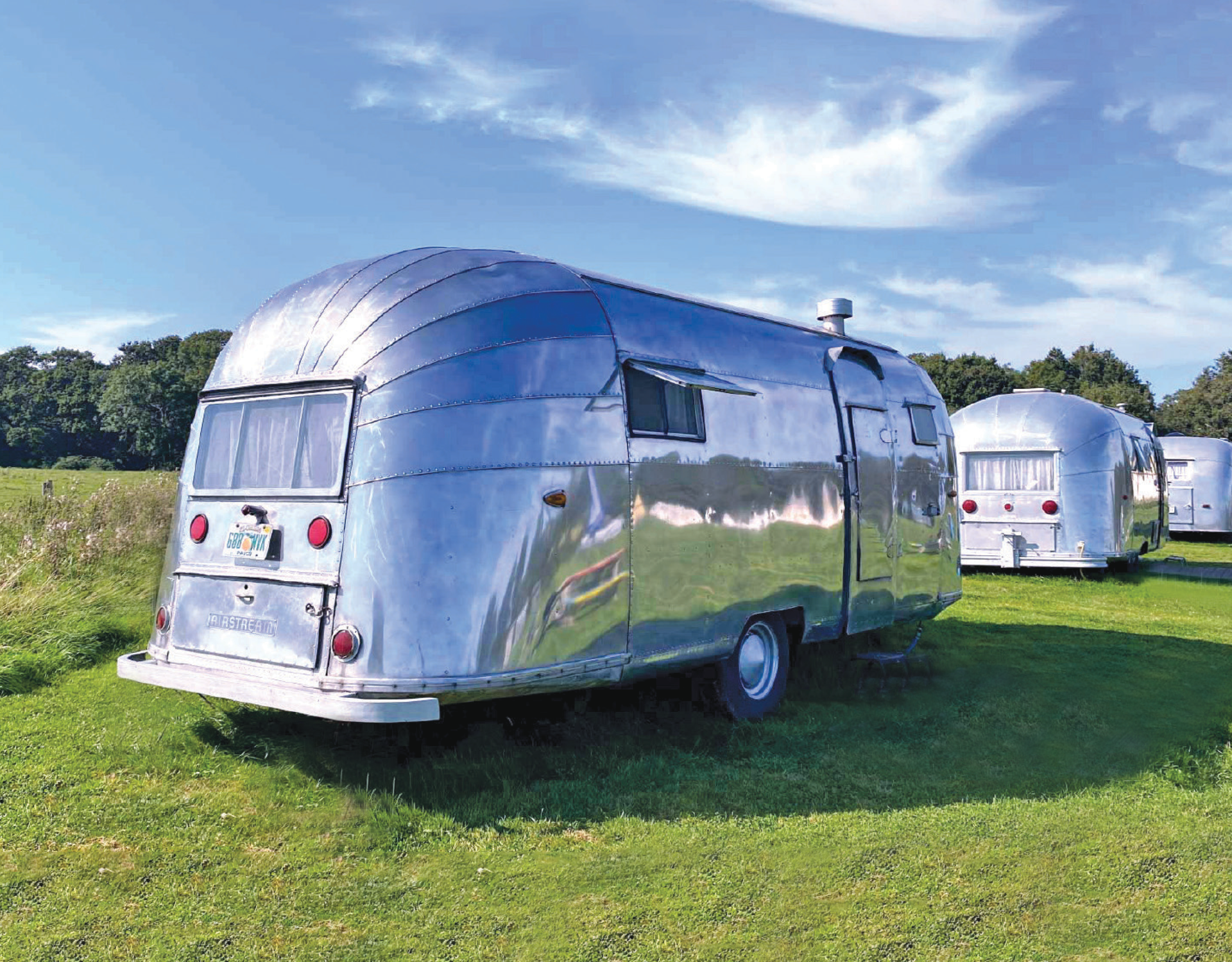 Shiny, silver, vintage travel trailers parked on a grassy field under a blue sky.