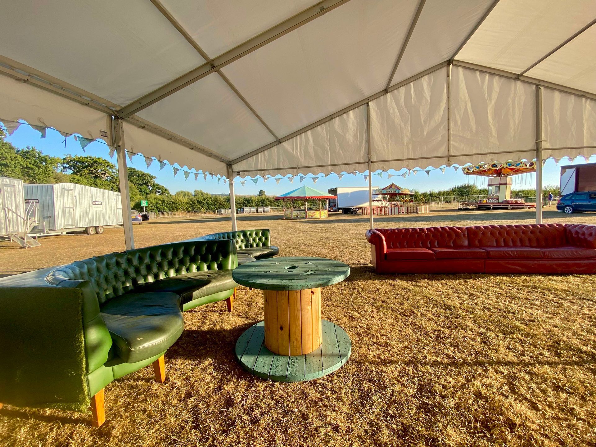 Green and red leather couches under a tent with a spool table, fairground in the background.