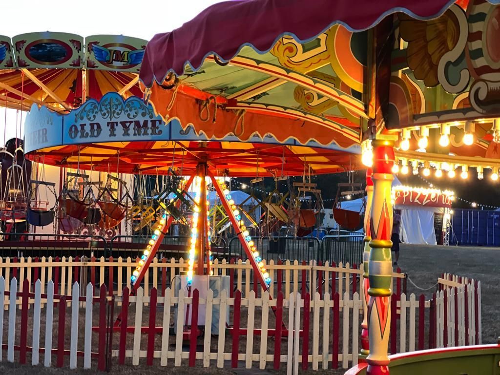 Carousel ride with brightly lit canopy and small fence; other carnival rides in background.