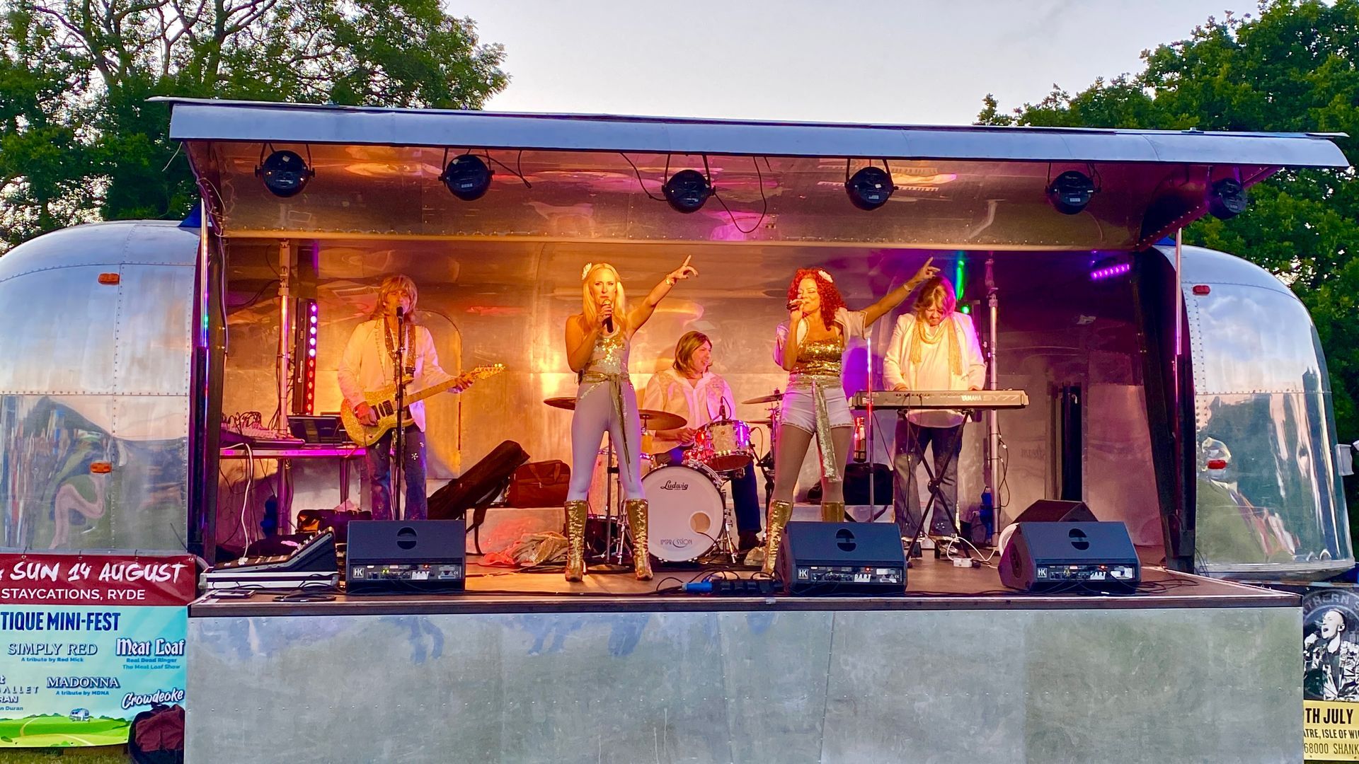 Band performing on a stage built from a silver trailer. Musicians play instruments, sing, under stage lights.