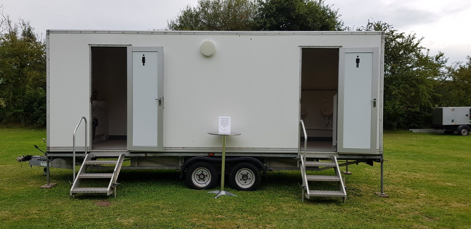 White portable restroom trailer on grass with open doors and steps.