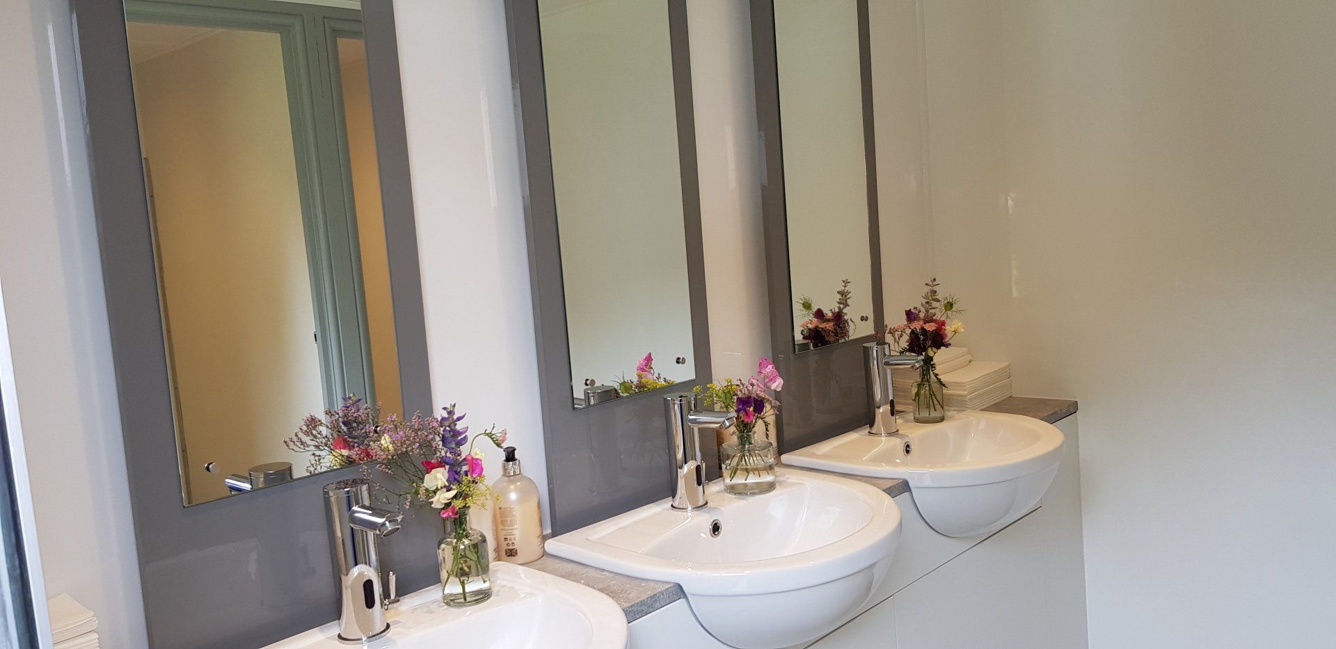 Three sinks with mirrors, small flower arrangements, and soap bottles in a restroom. White walls and gray mirror frames.