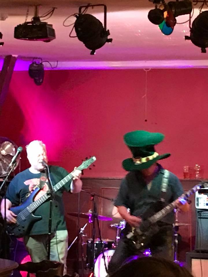 Two guitarists on stage, one wearing a large green hat, playing music in a dimly lit bar with pink and white lights.