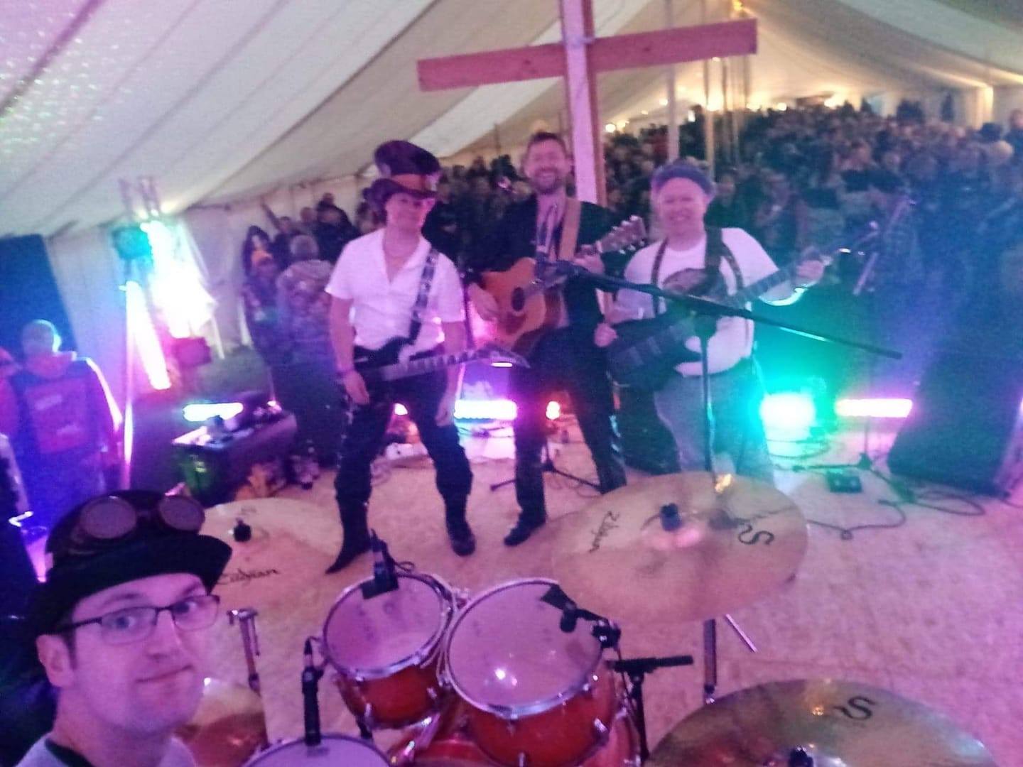 Band performing on stage in a tent, crowd in background. Musicians play guitar, drums, and sing. A cross is behind the stage.