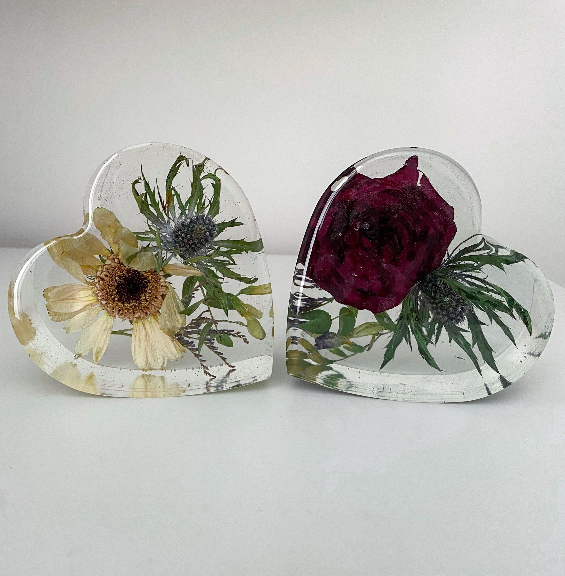 Two heart-shaped resin paperweights with preserved flowers inside. One has a yellow daisy, the other a deep red rose.
