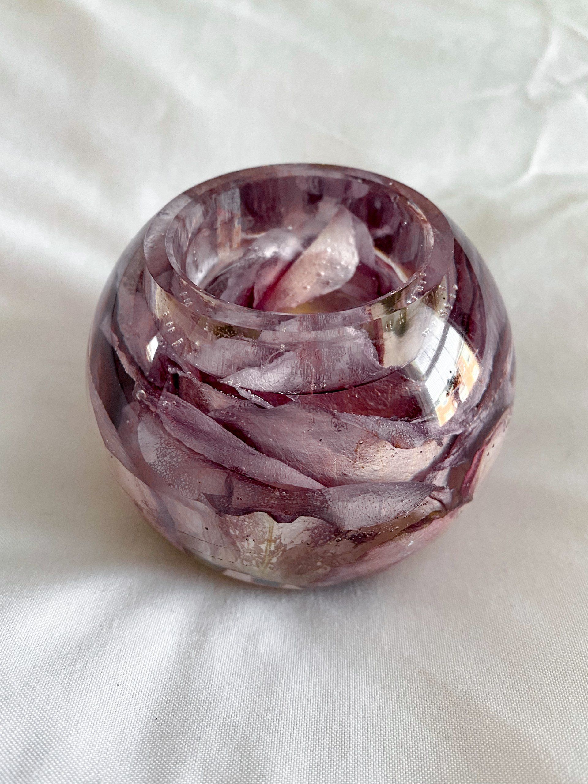 Round resin candle holder with dried purple petals, resting on white fabric.