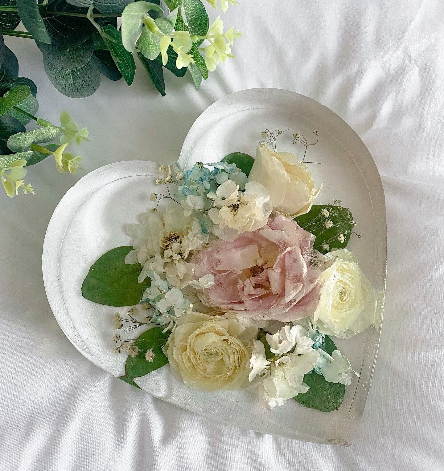Heart-shaped clear resin paperweight with dried flowers. Roses, hydrangeas, and leaves in pale pink, blue, cream, and green.