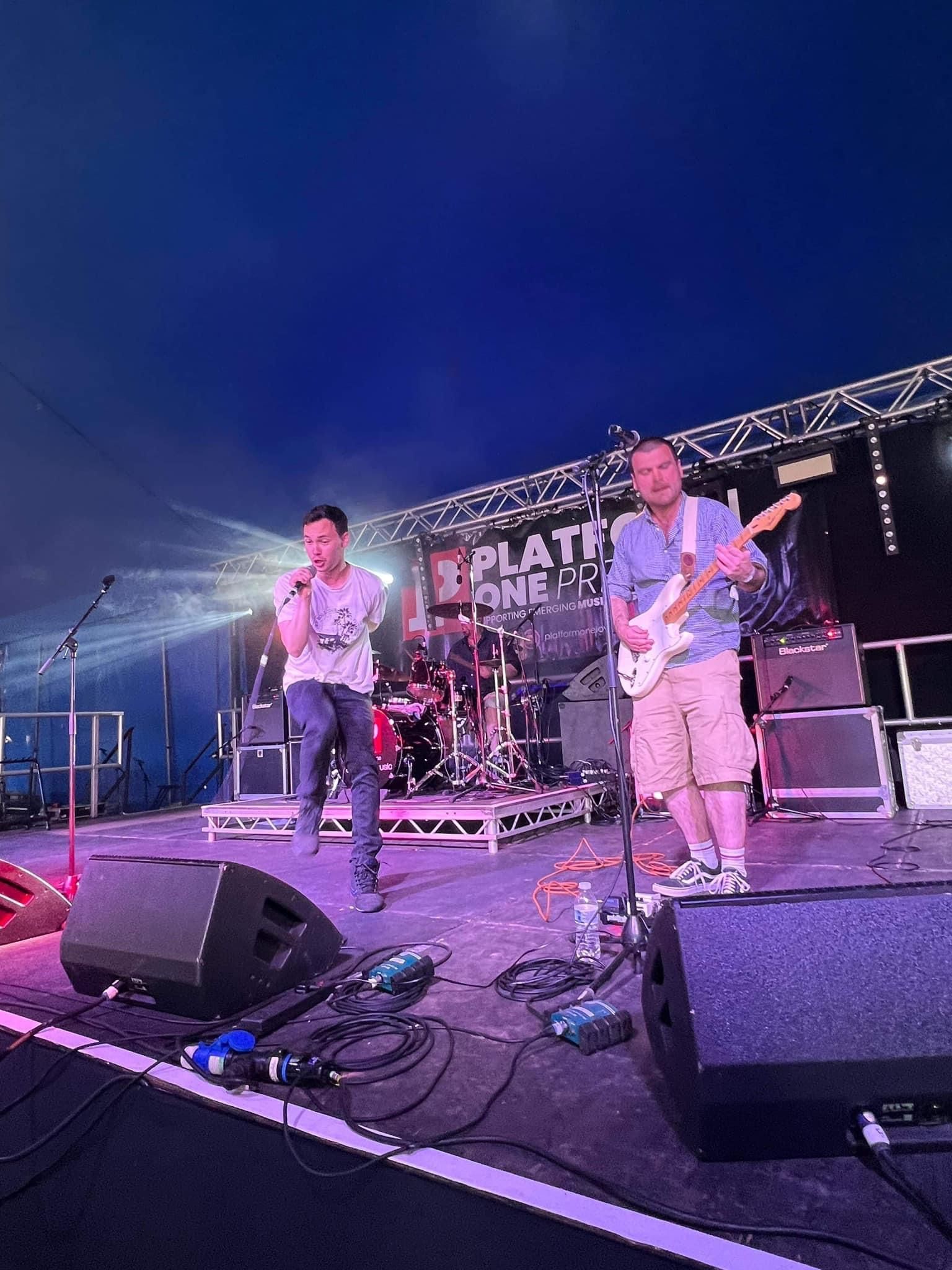 Band performing on stage, singer with microphone, guitarist playing, drums in background, outdoor setting.