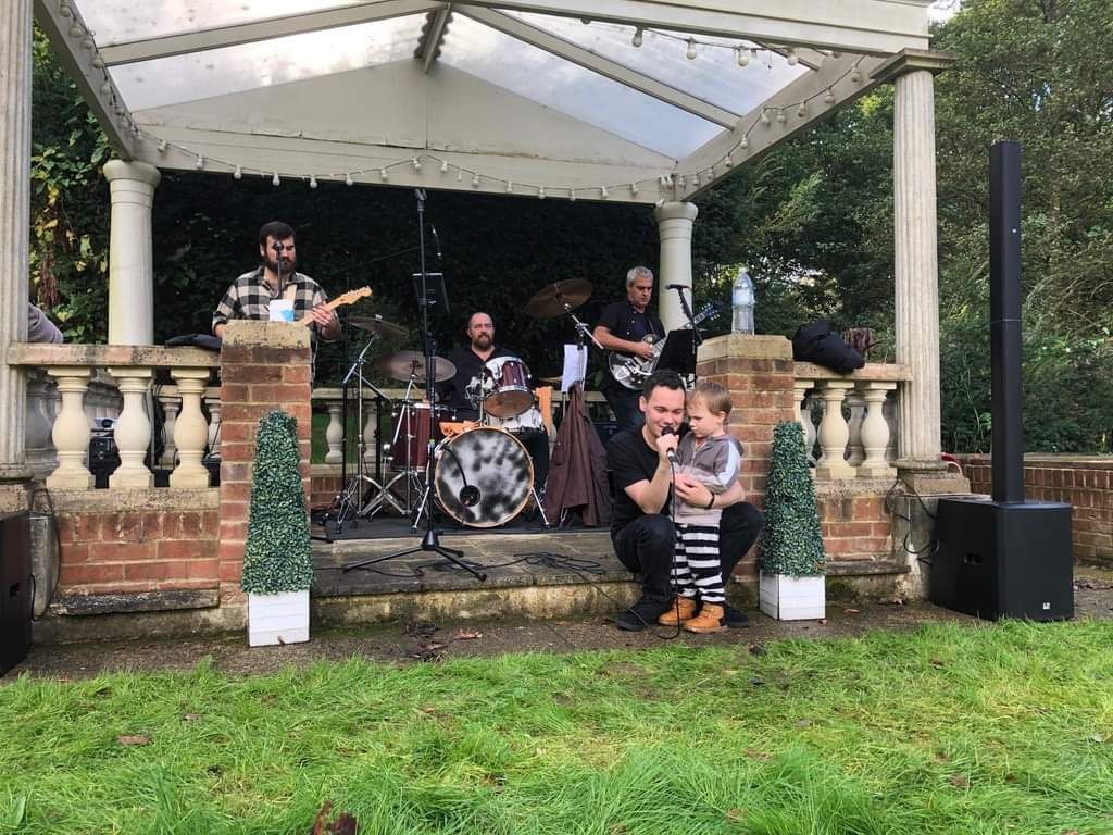 Band playing under a gazebo; man holding a child sings into a microphone.