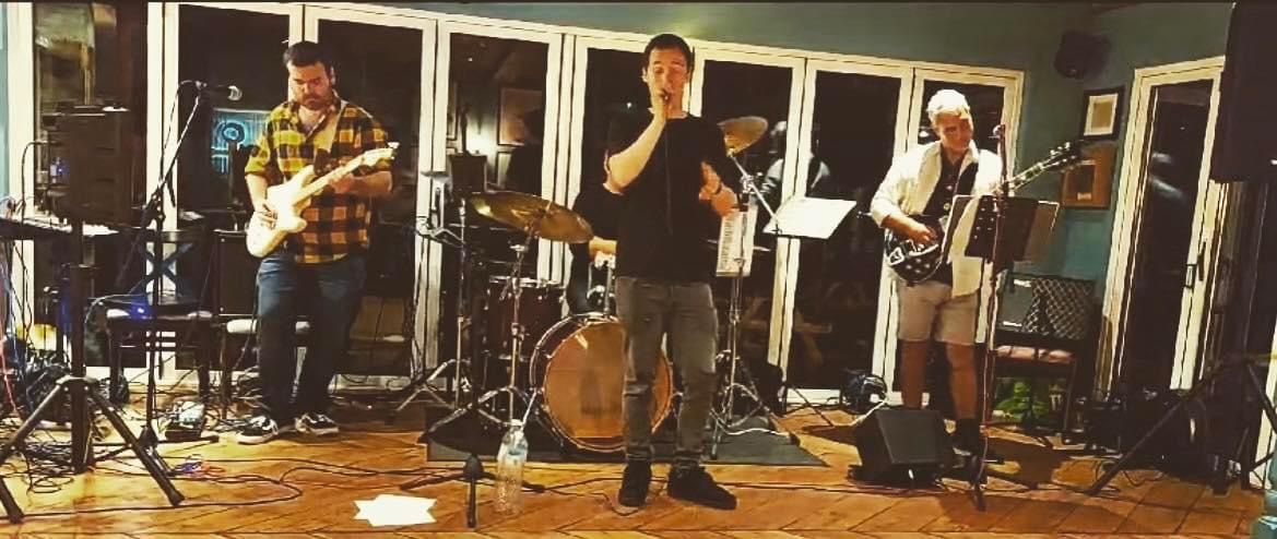 A band of four playing music in a room with large windows. The lead singer is in the center with two guitarists on each side and a drummer.