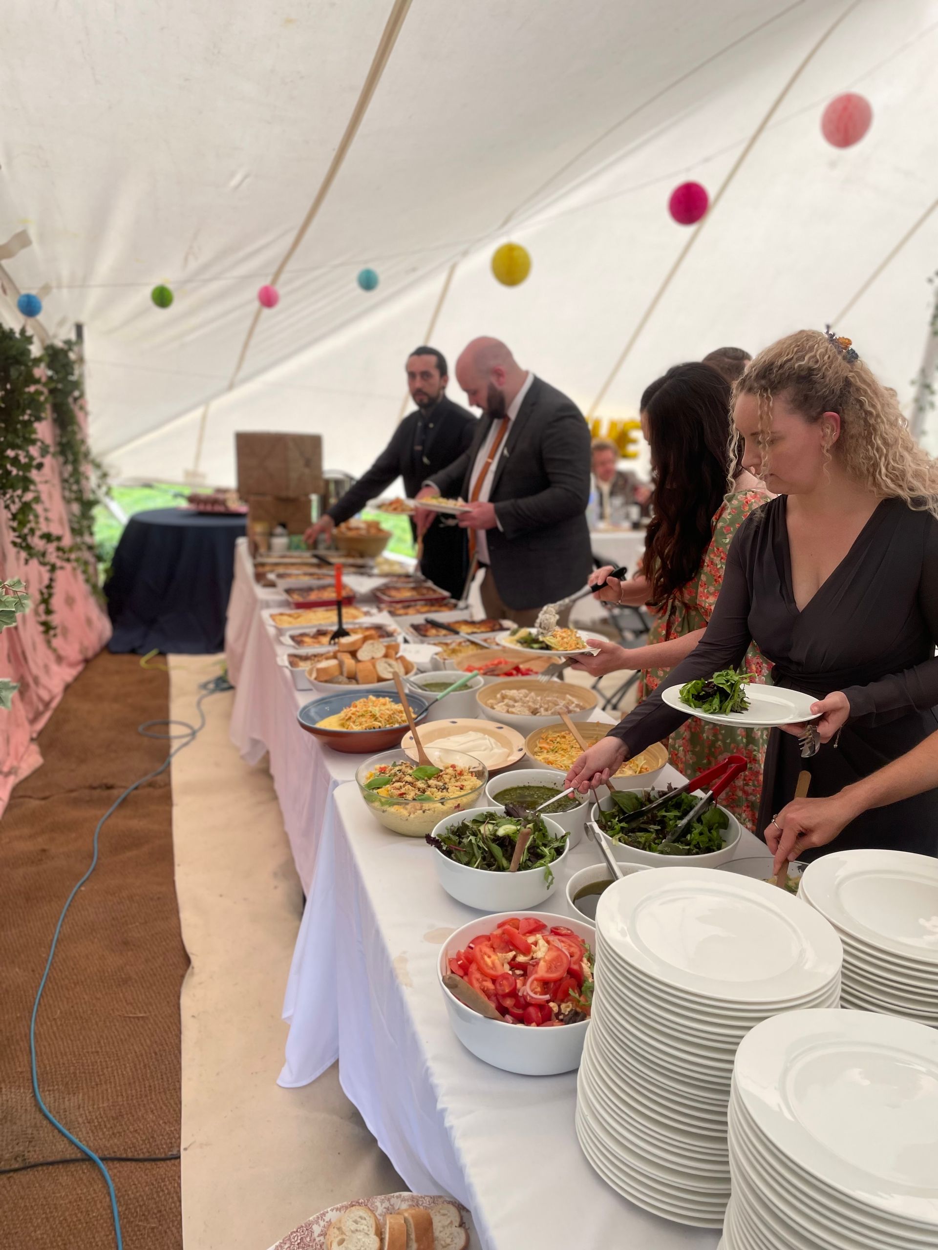 People serving themselves from a buffet table under a white tent; various food items displayed.