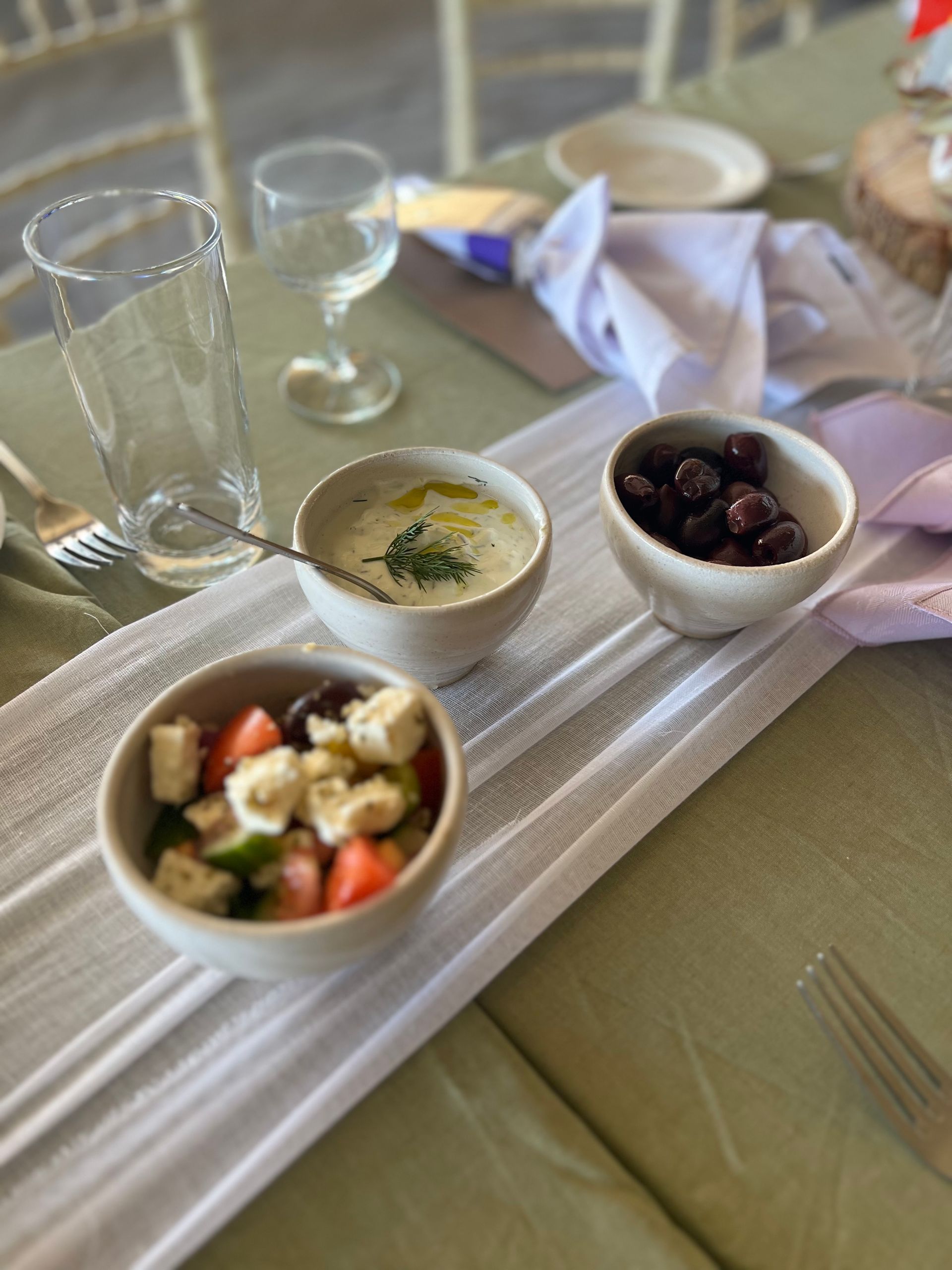 Bowls of Greek salad, olives, and dip on a table with a water glass and napkin.