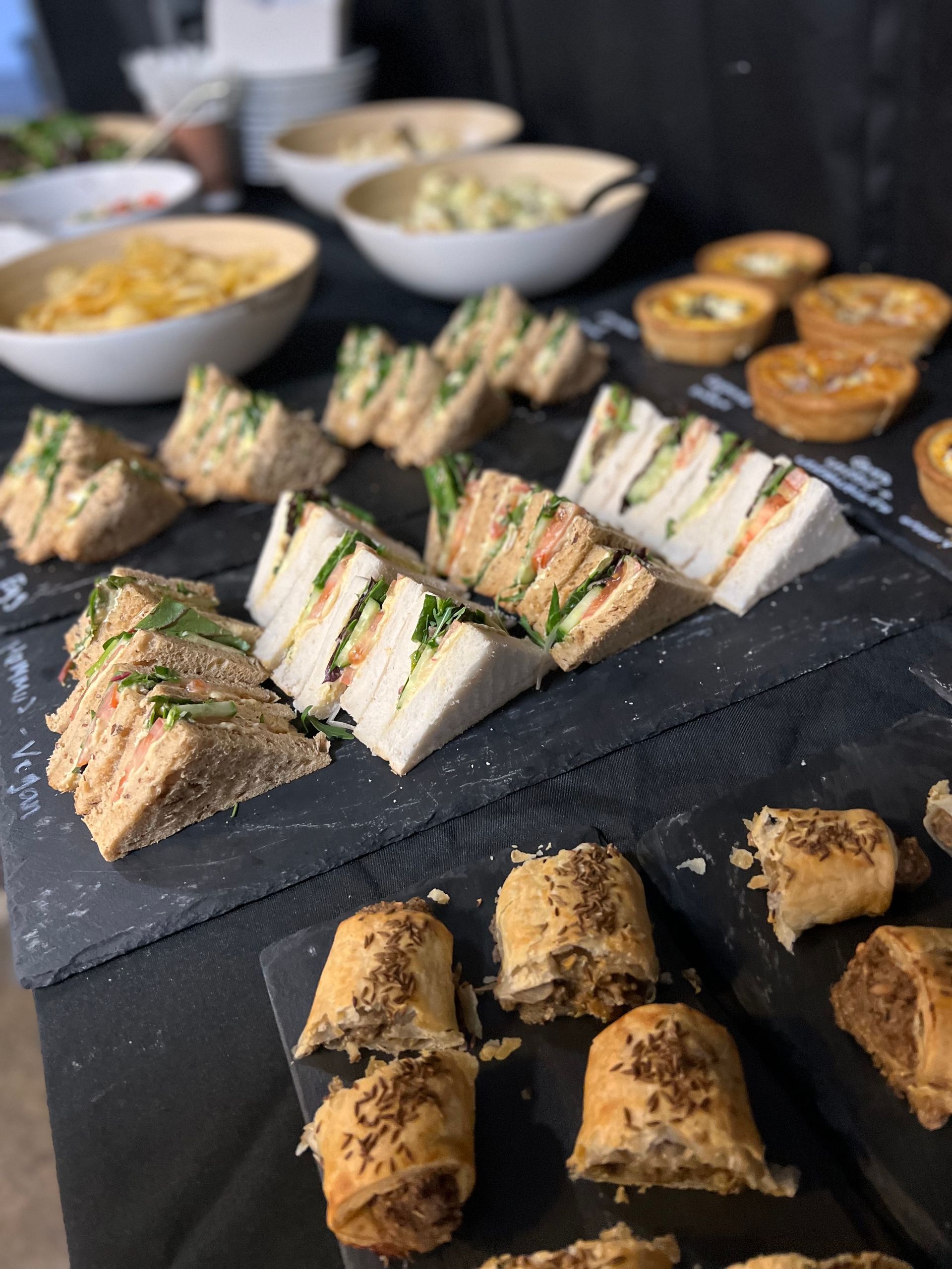 Sandwiches, tarts, and sausage rolls arranged on a table with bowls of snacks in the background.