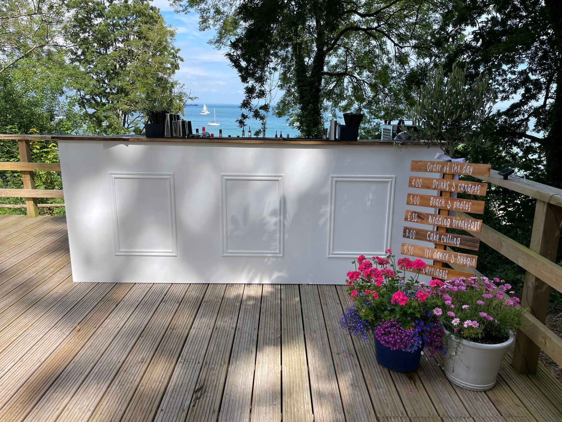 White bar decorated with framed panels, flowers, and a sign on a wooden deck overlooking water.