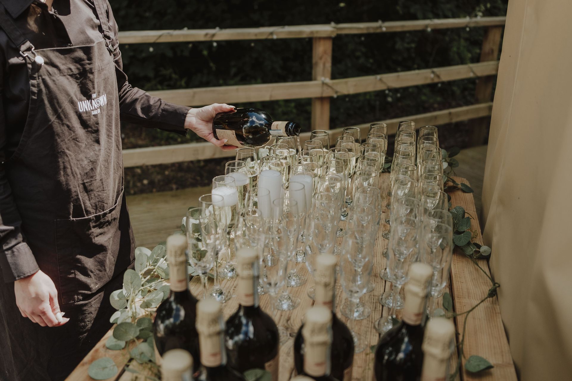 Bartender pouring wine into champagne flutes on a wooden table. Bottles and greenery are also present.