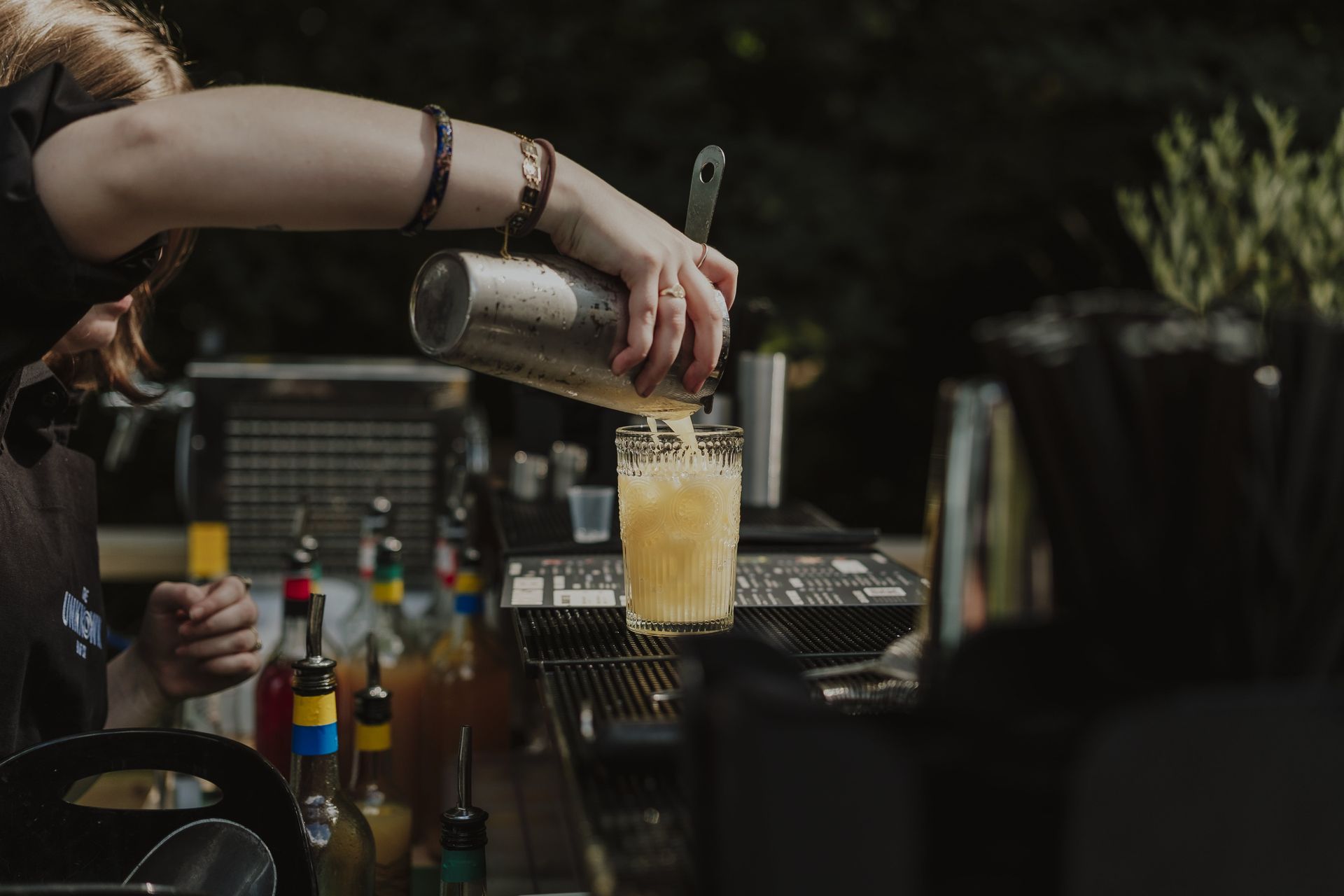 Bartender pouring a yellow drink from a shaker into a glass, outdoor bar setting.