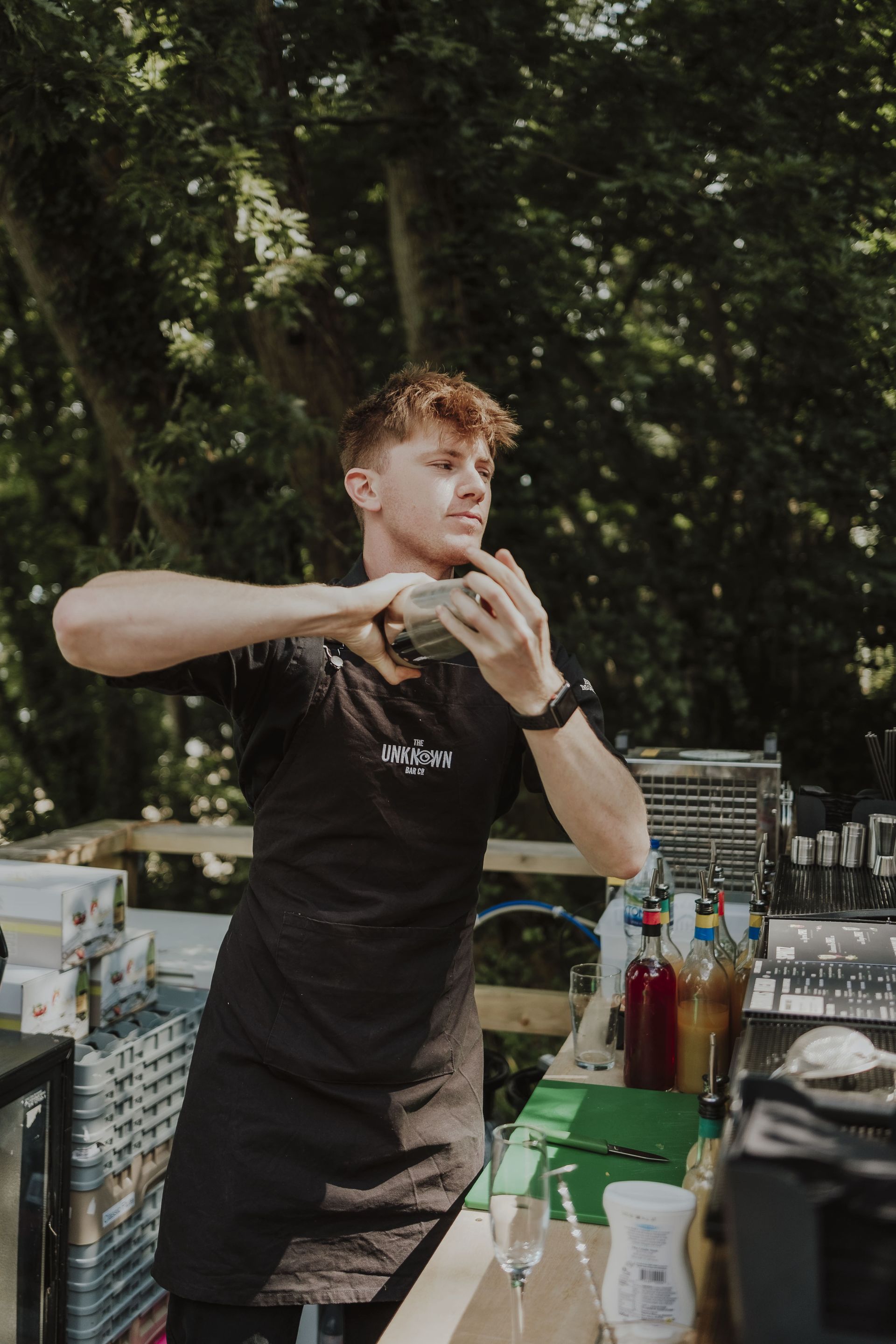 Bartender shaking a cocktail at an outdoor bar; mixing drinks, green and red bottles visible.