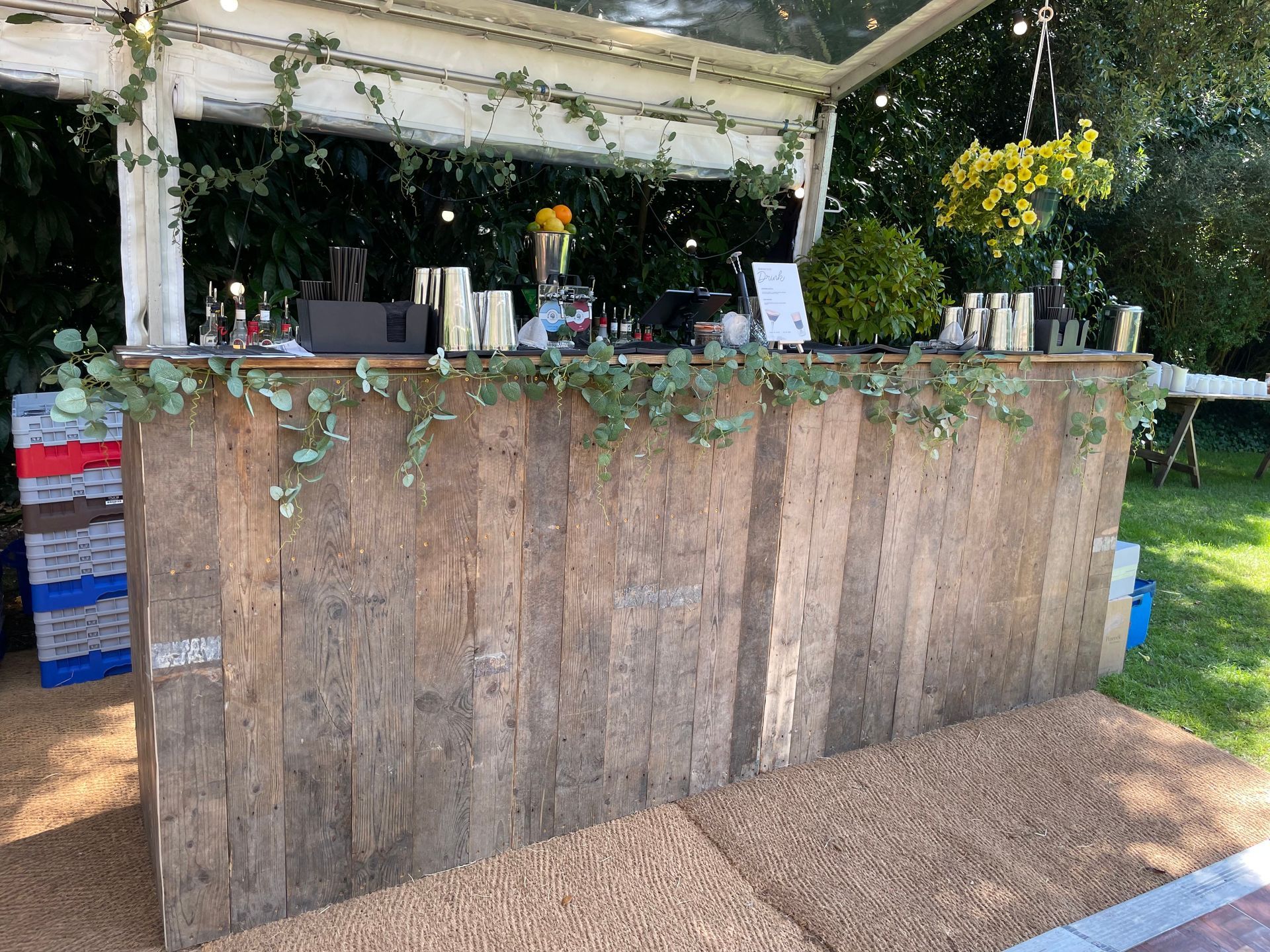 Rustic wooden bar outdoors, adorned with greenery, set up for an event.