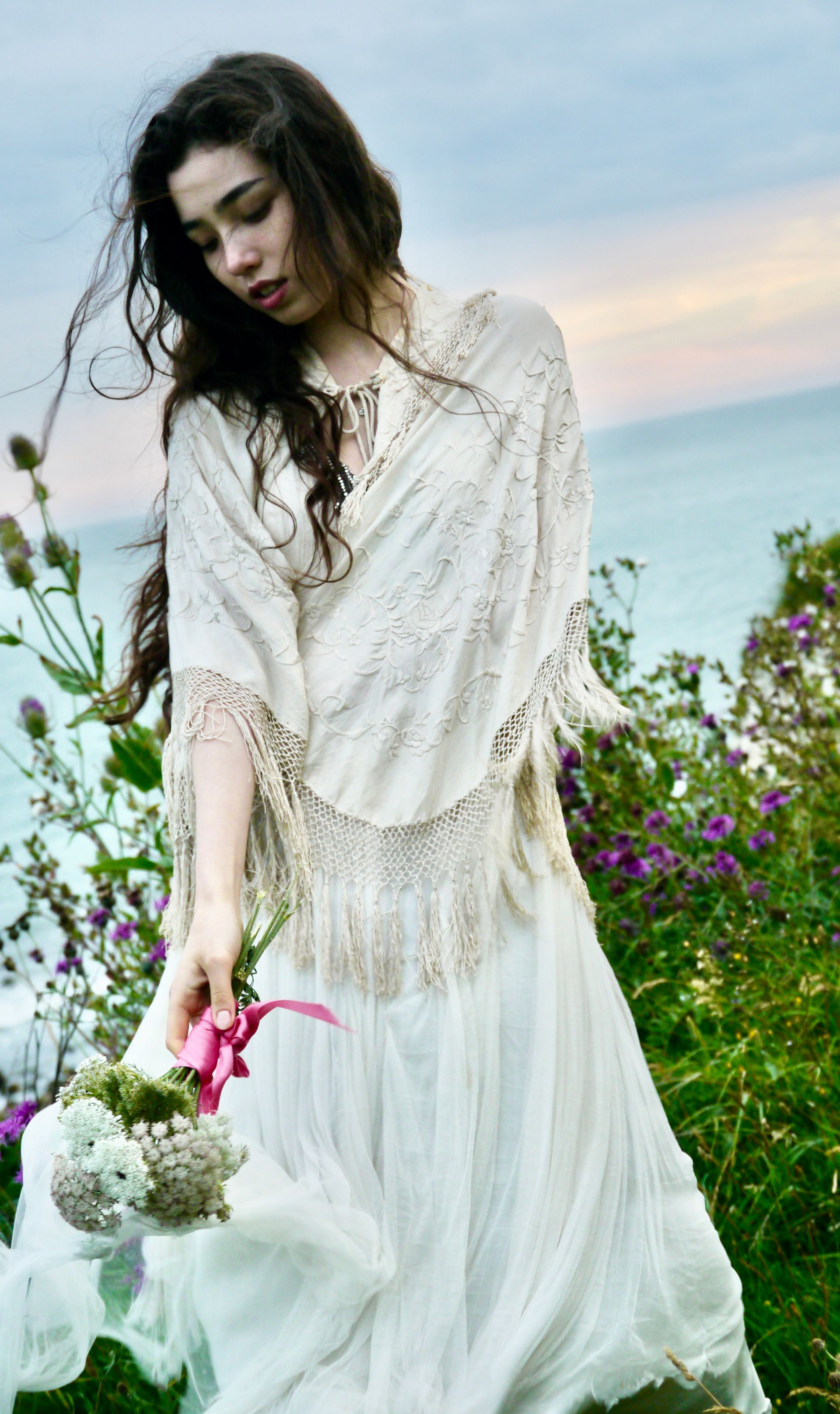Woman in white dress and shawl holding flowers by the sea.