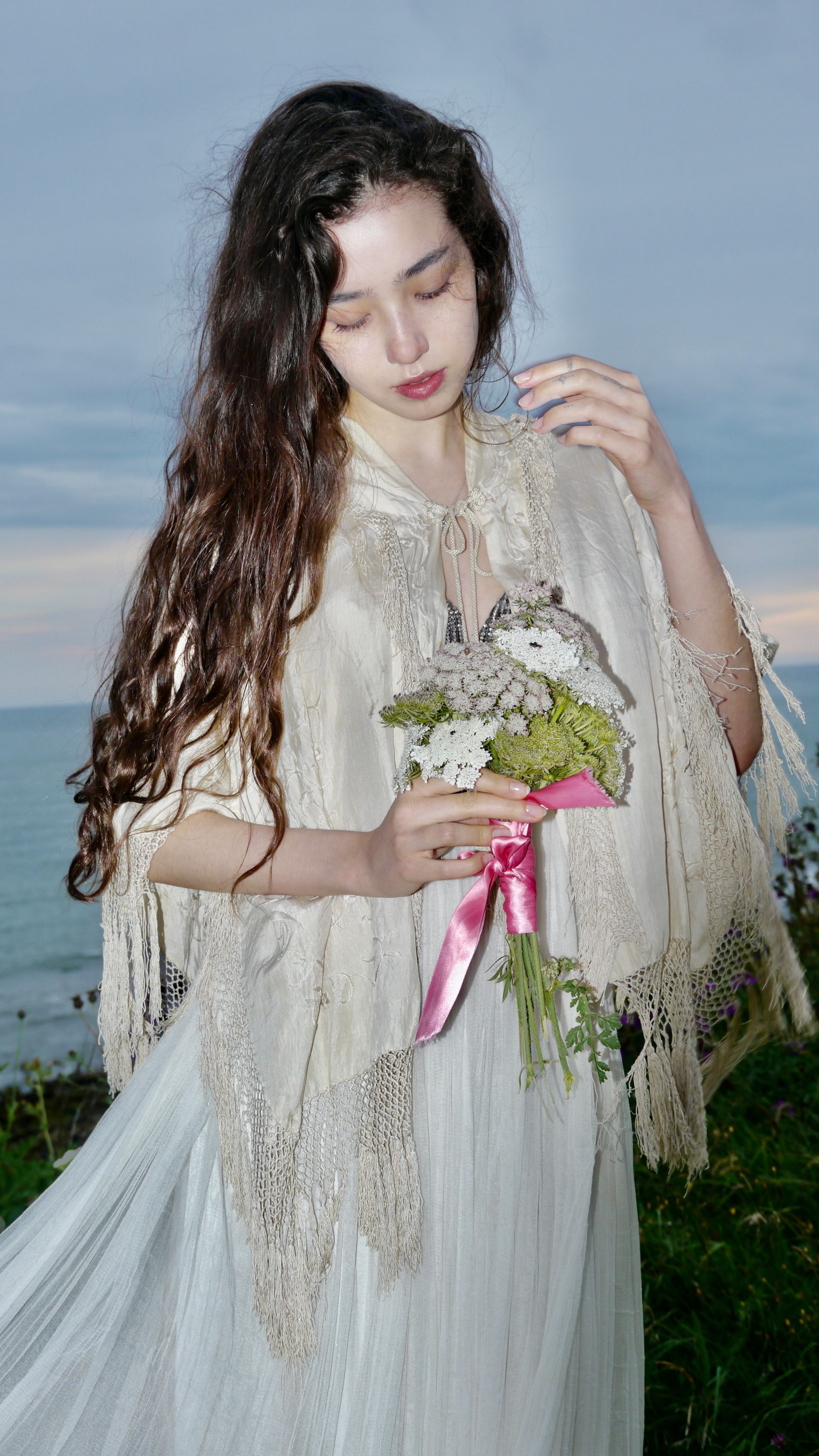 Woman in white lace shawl and dress holding flowers, standing by the sea.