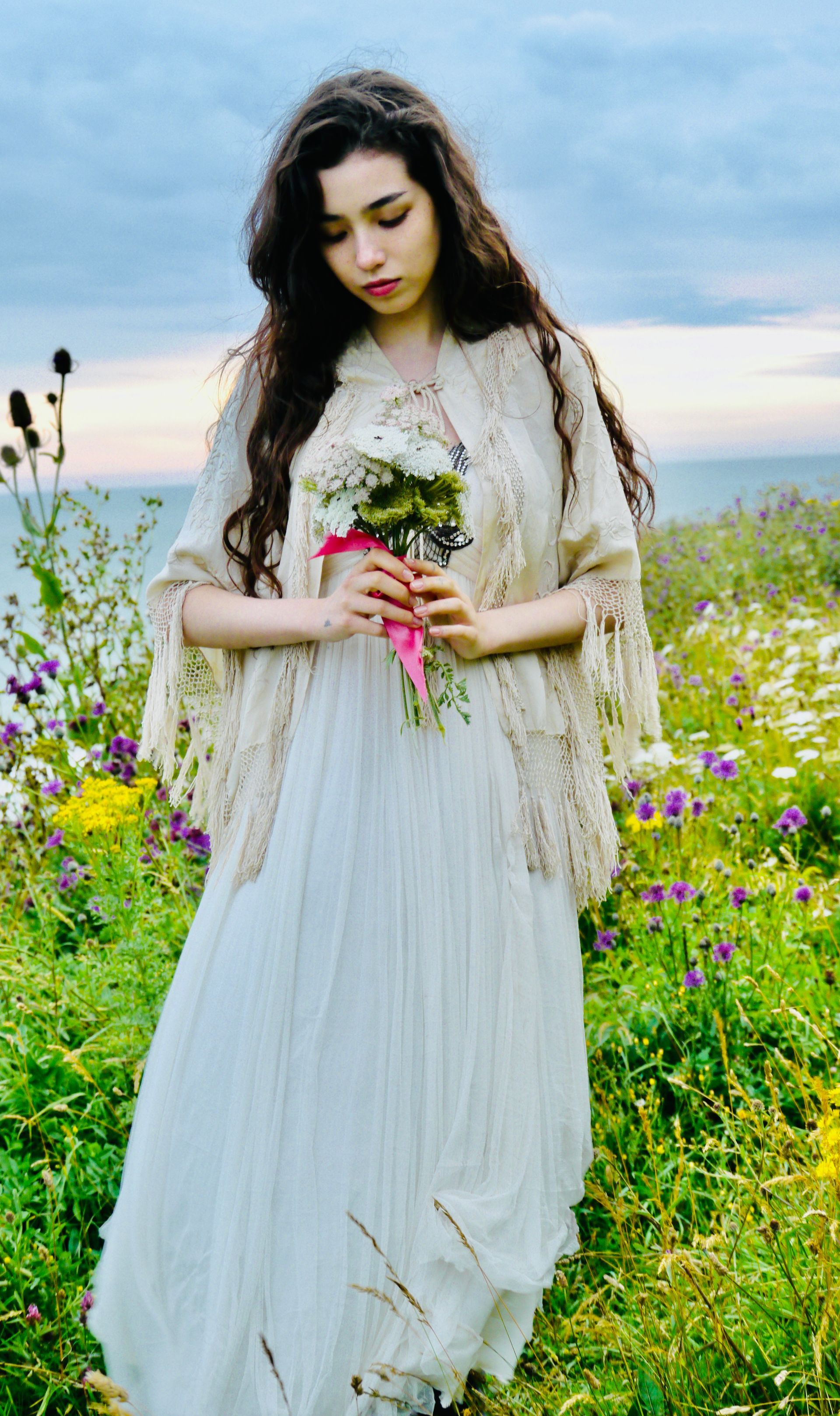 Woman in a white dress holds flowers in a field of wildflowers, wearing a shawl, looking down.