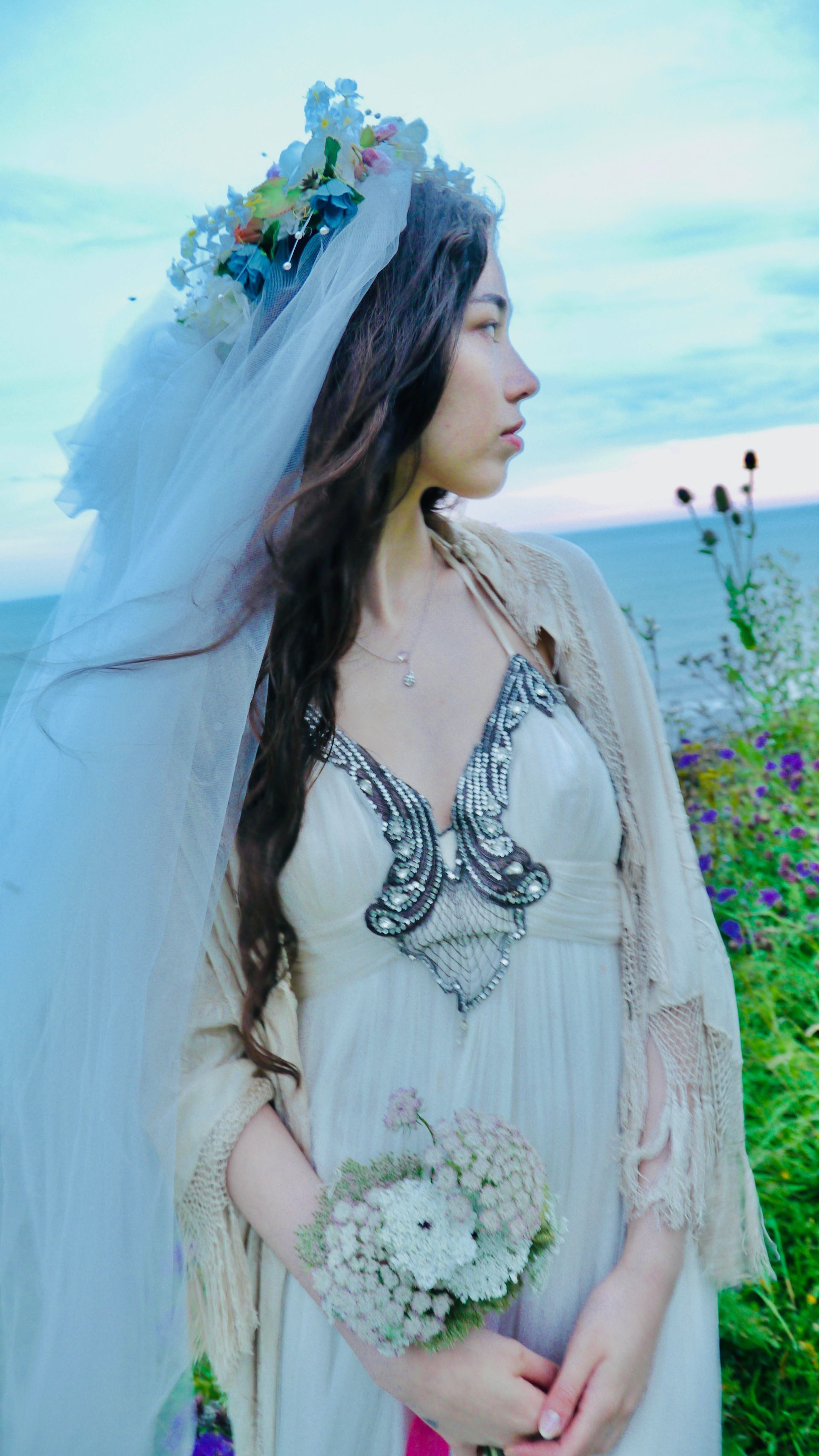 Woman wearing a flower crown and veil, holding bouquet, looking toward the ocean.