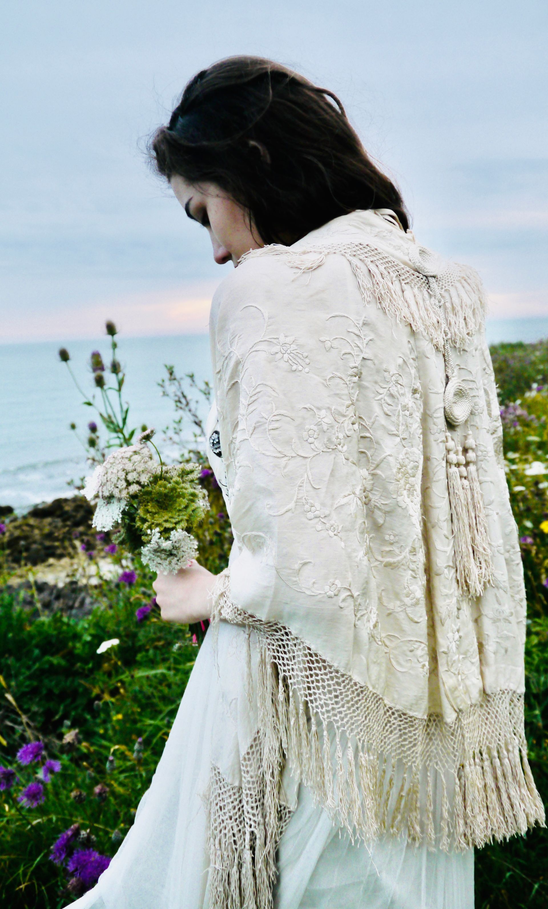 Woman in white dress and shawl holding flowers, standing near a body of water, facing away.