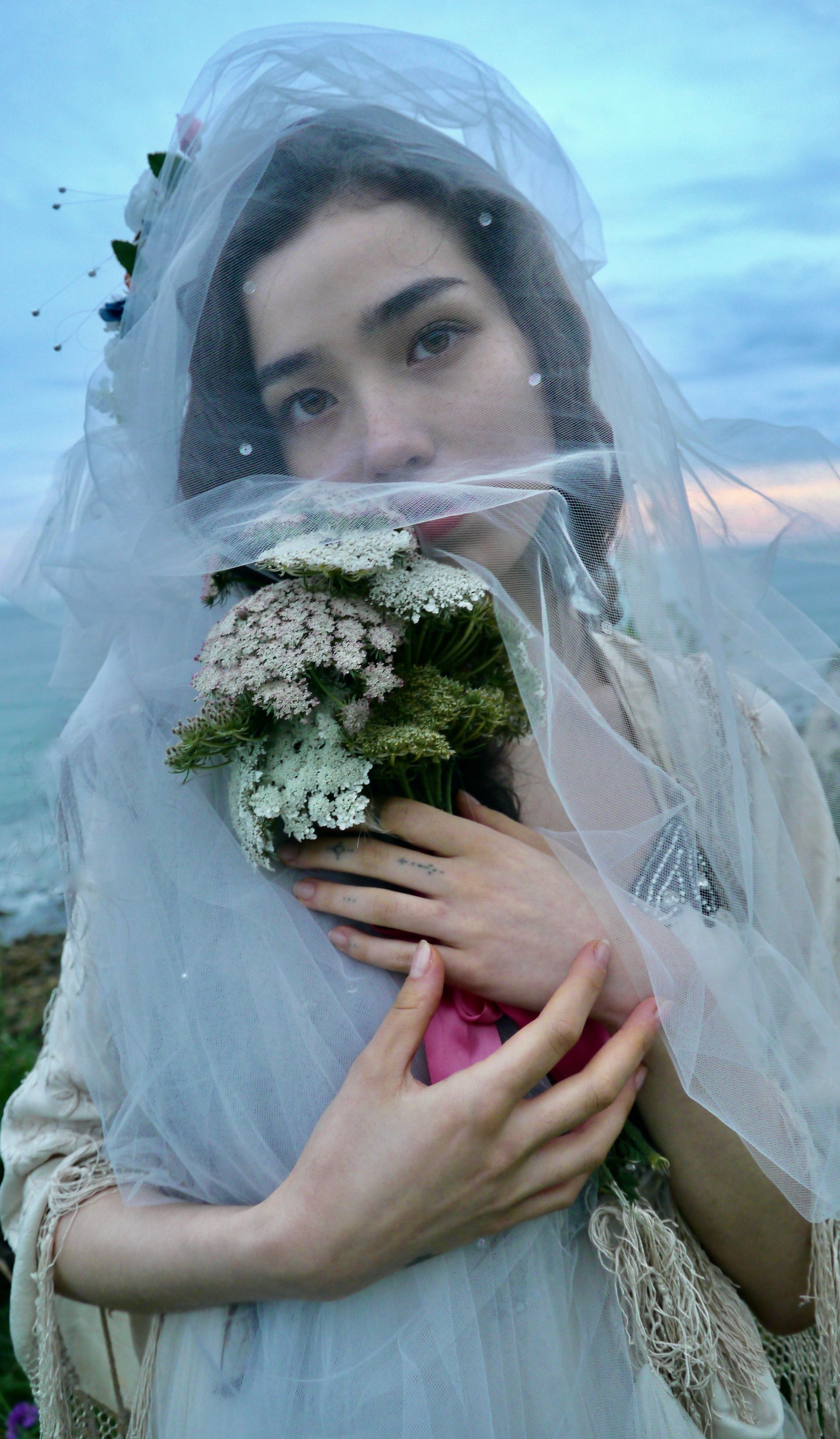 Woman with veil, holding flowers, looking at camera by the sea.