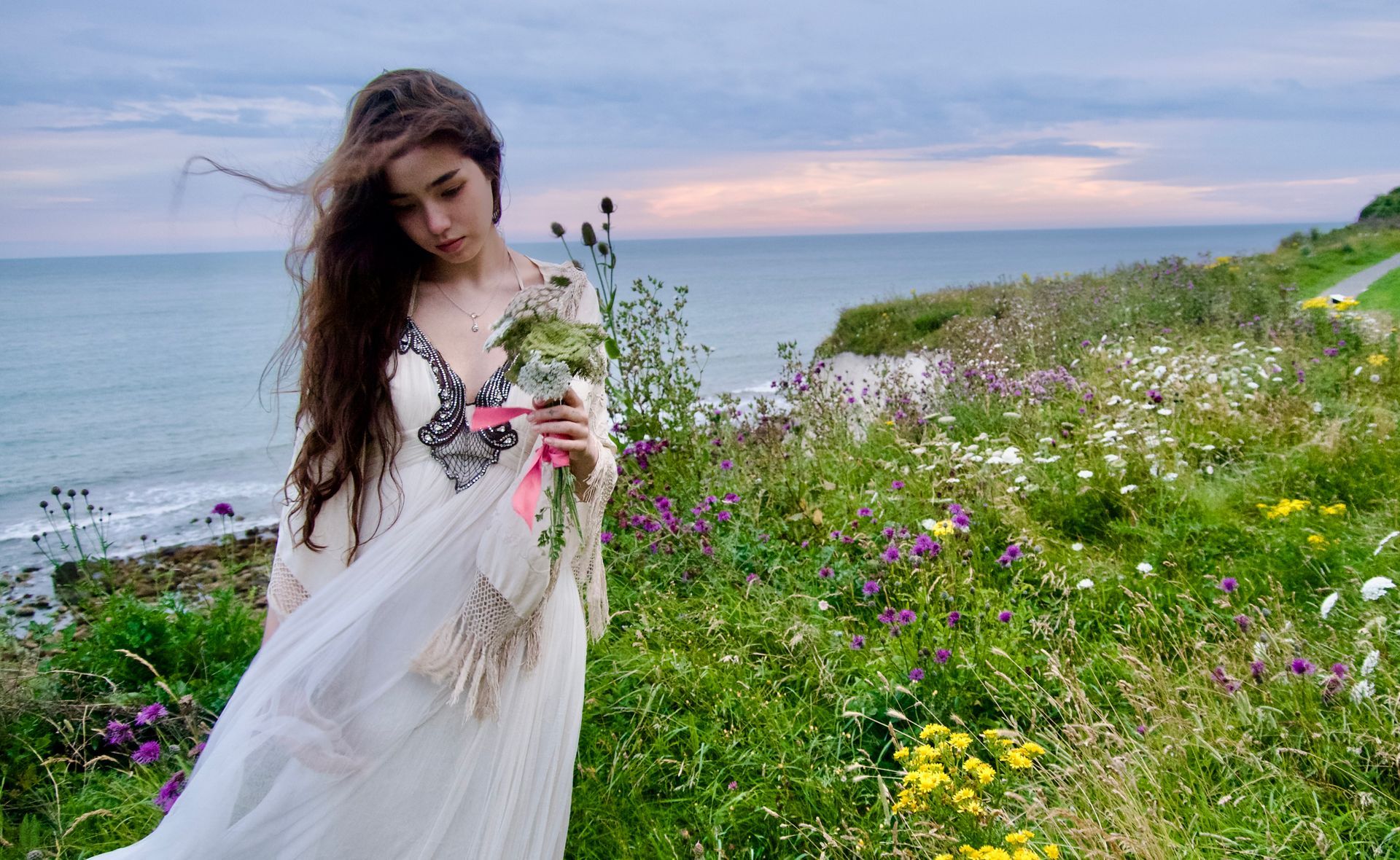 Woman in white dress with flowers, standing by the ocean with a cloudy sky.