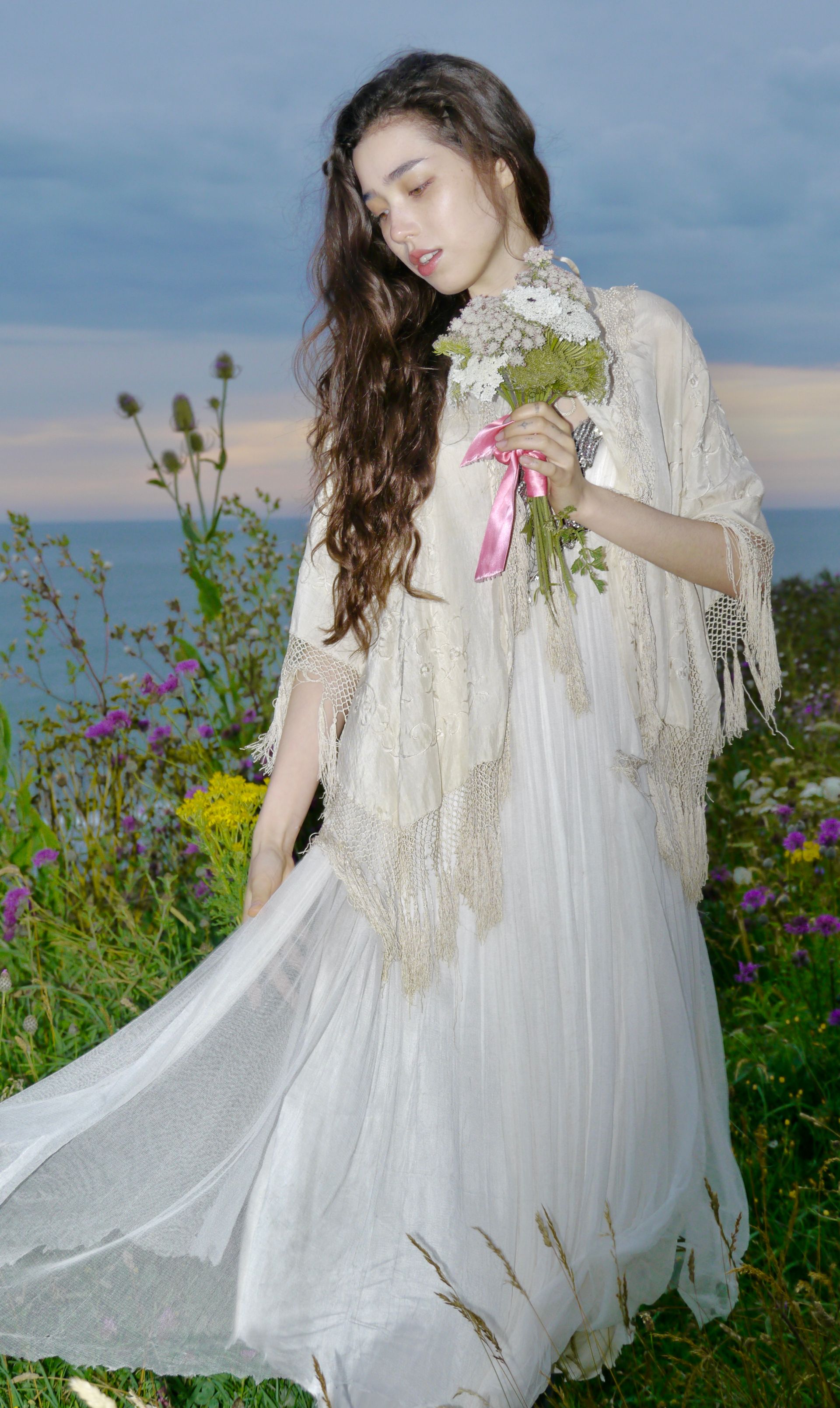 Woman in white dress and shawl holding flowers, standing near a body of water and flowers.