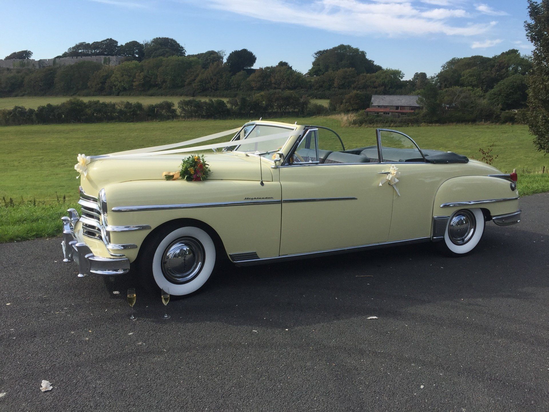 Cream-colored vintage convertible decorated with flowers, parked on asphalt with a grassy backdrop.