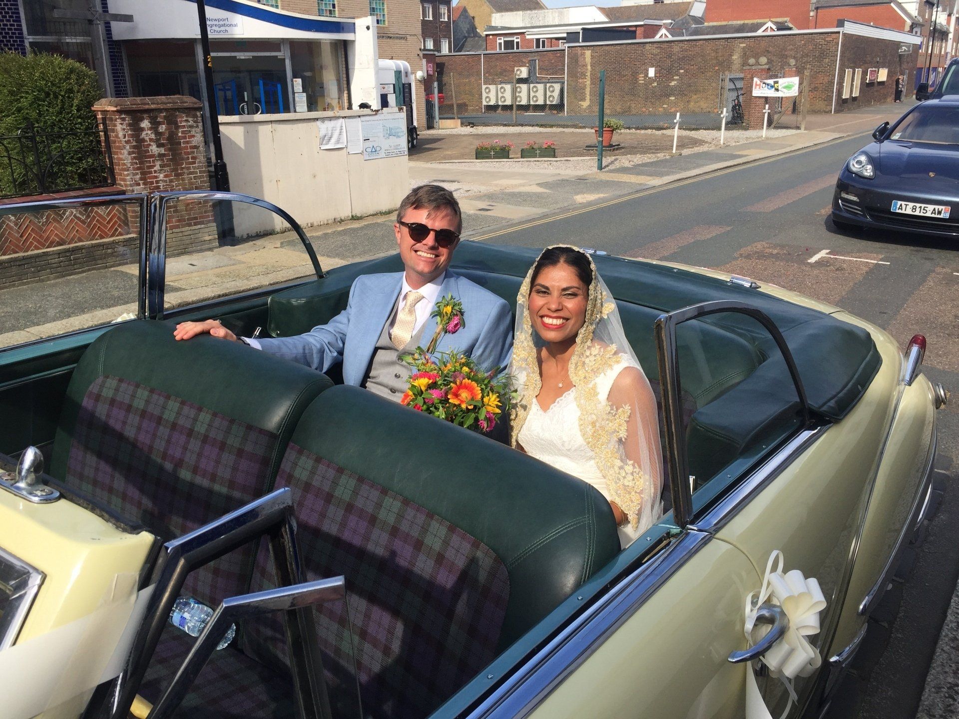 Newlyweds smiling in vintage convertible; bride in veil, groom in suit.