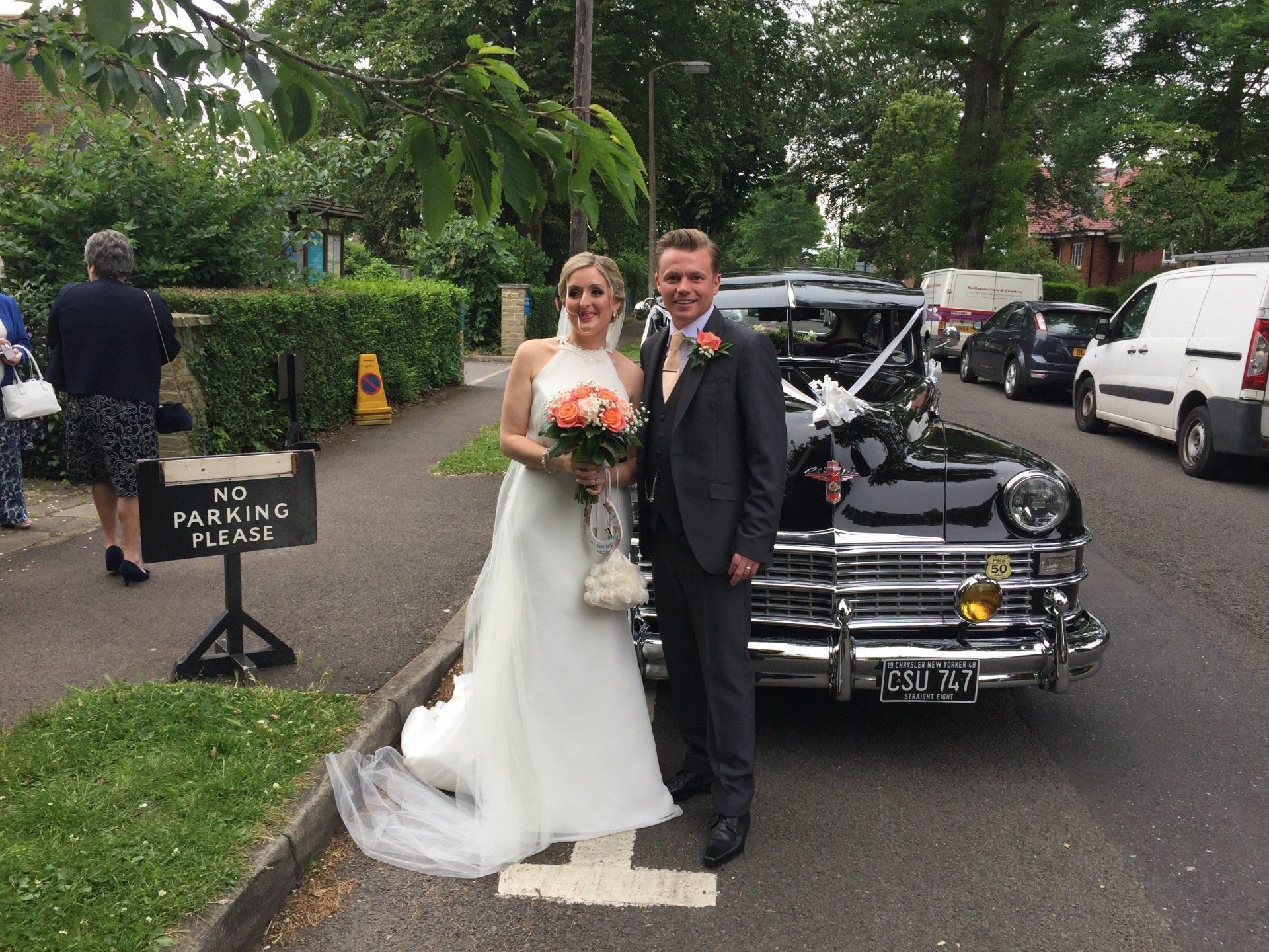 Bride and groom pose by a black vintage car after their wedding. The bride holds a bouquet.