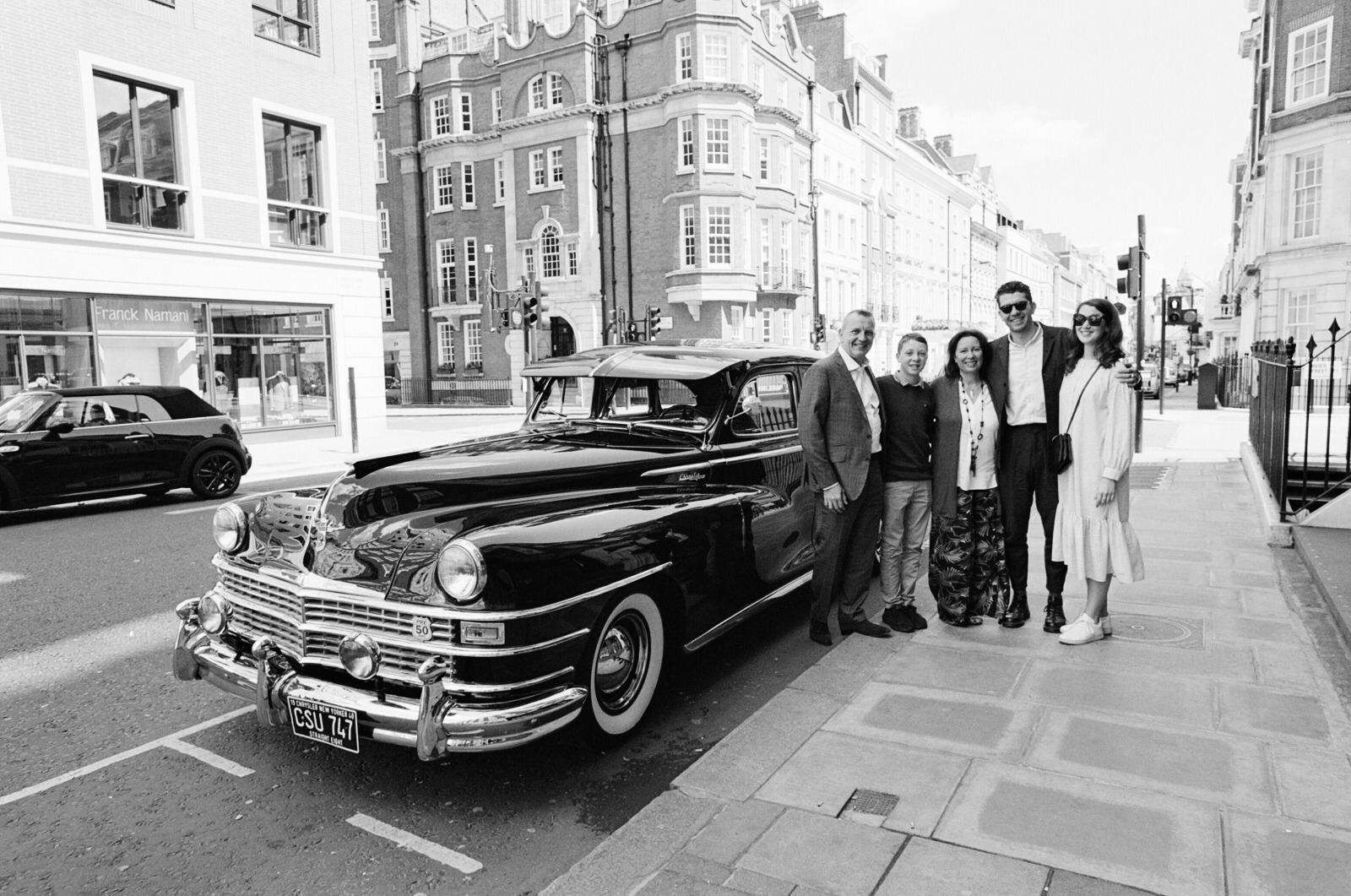 Group poses with a vintage black car on a city sidewalk. Buildings line the street.
