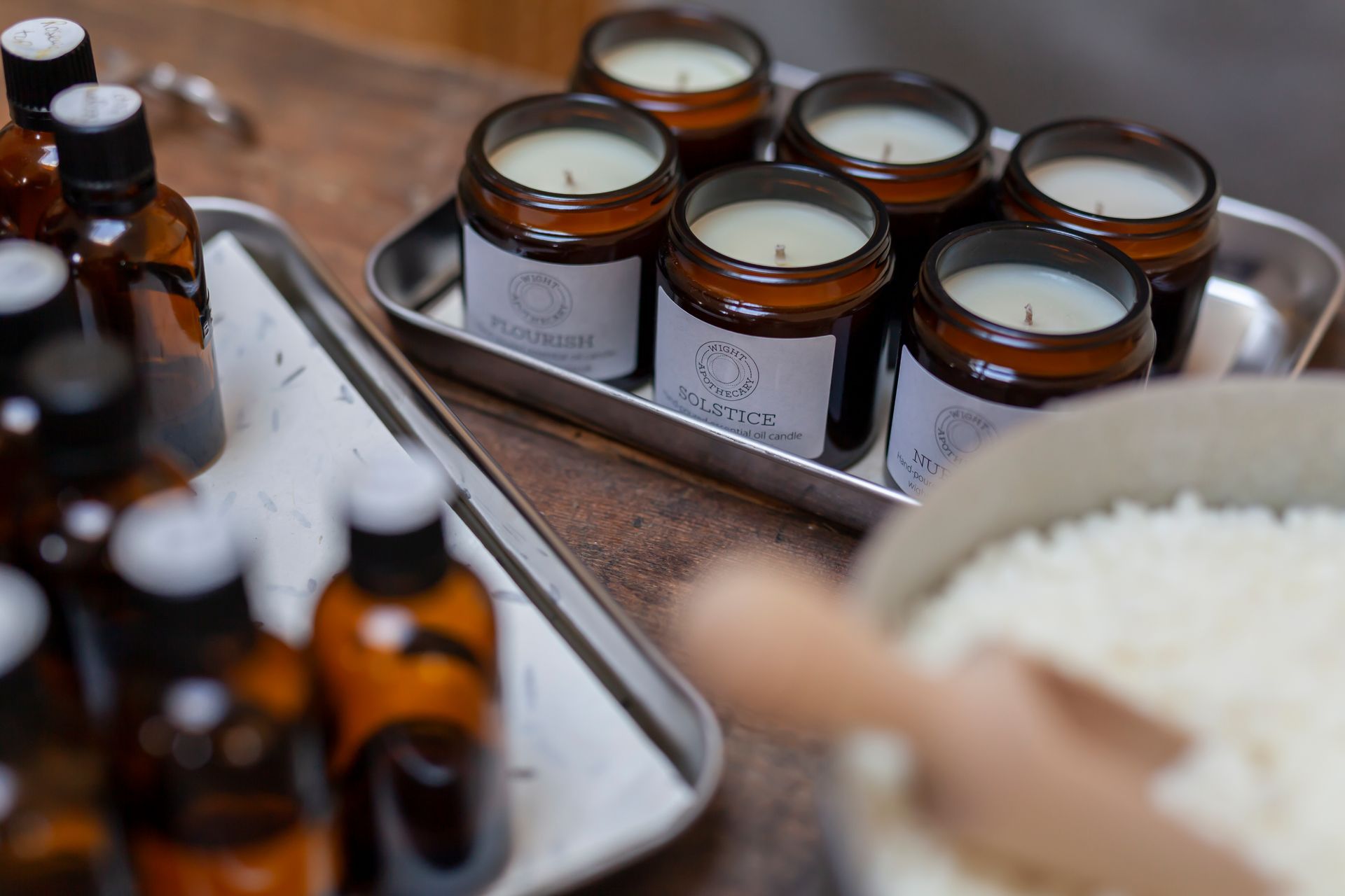 Candle-making setup: glass jars, brown bottles, wax, and a scoop on a wooden table.