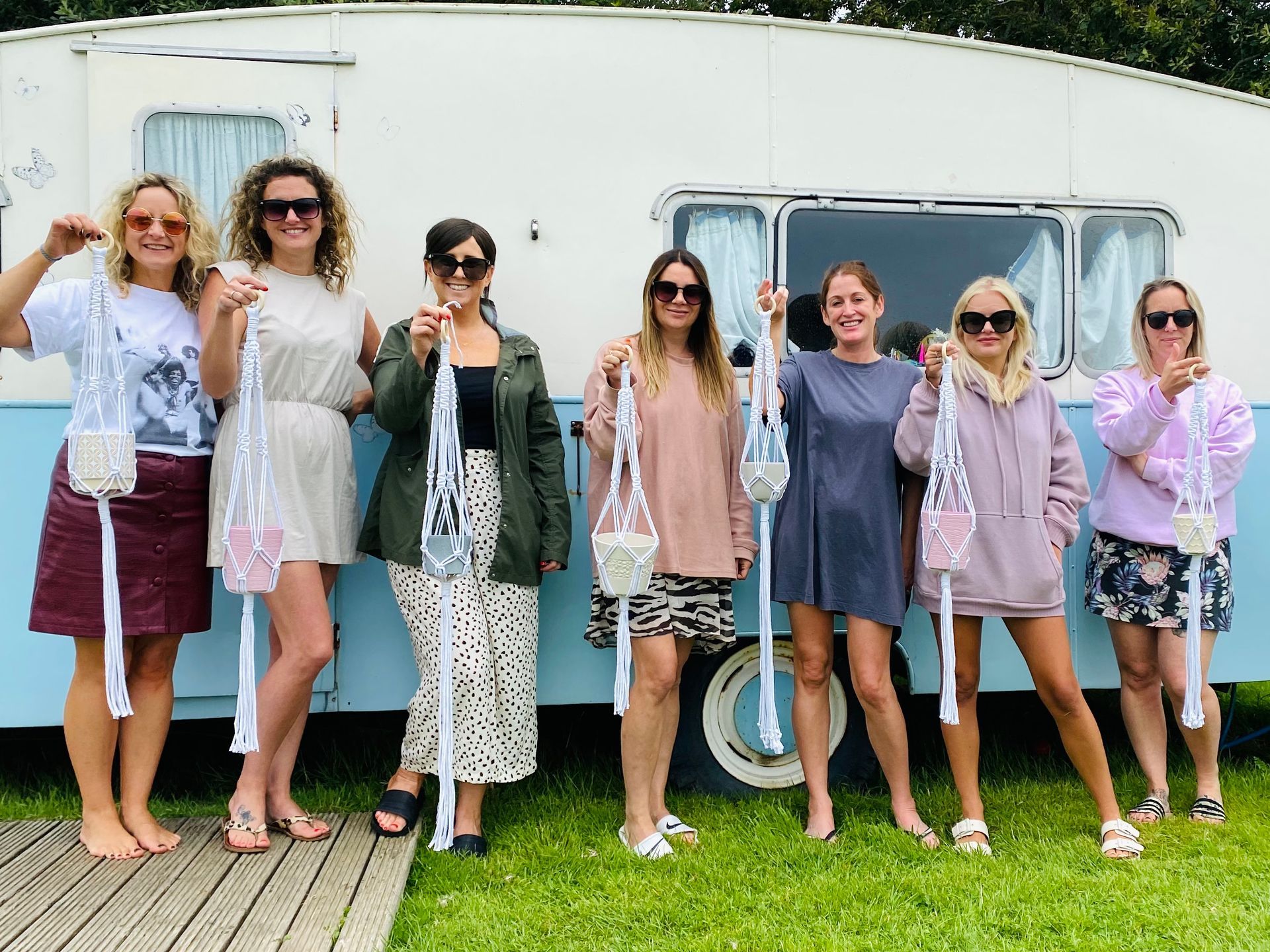 Seven women stand by a vintage camper, holding macrame plant hangers they made.