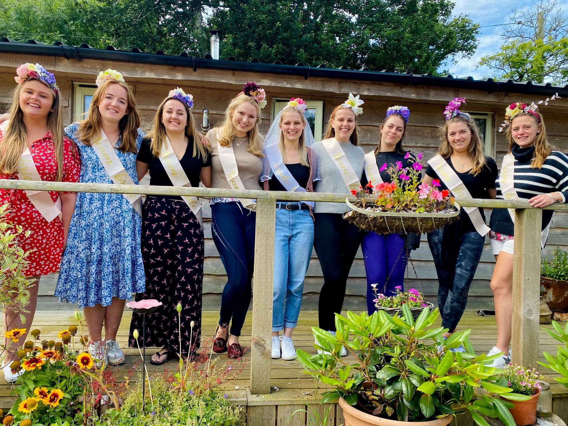 Group of women wearing flower crowns and sashes, posing in front of a wooden building, possibly a bridal party.