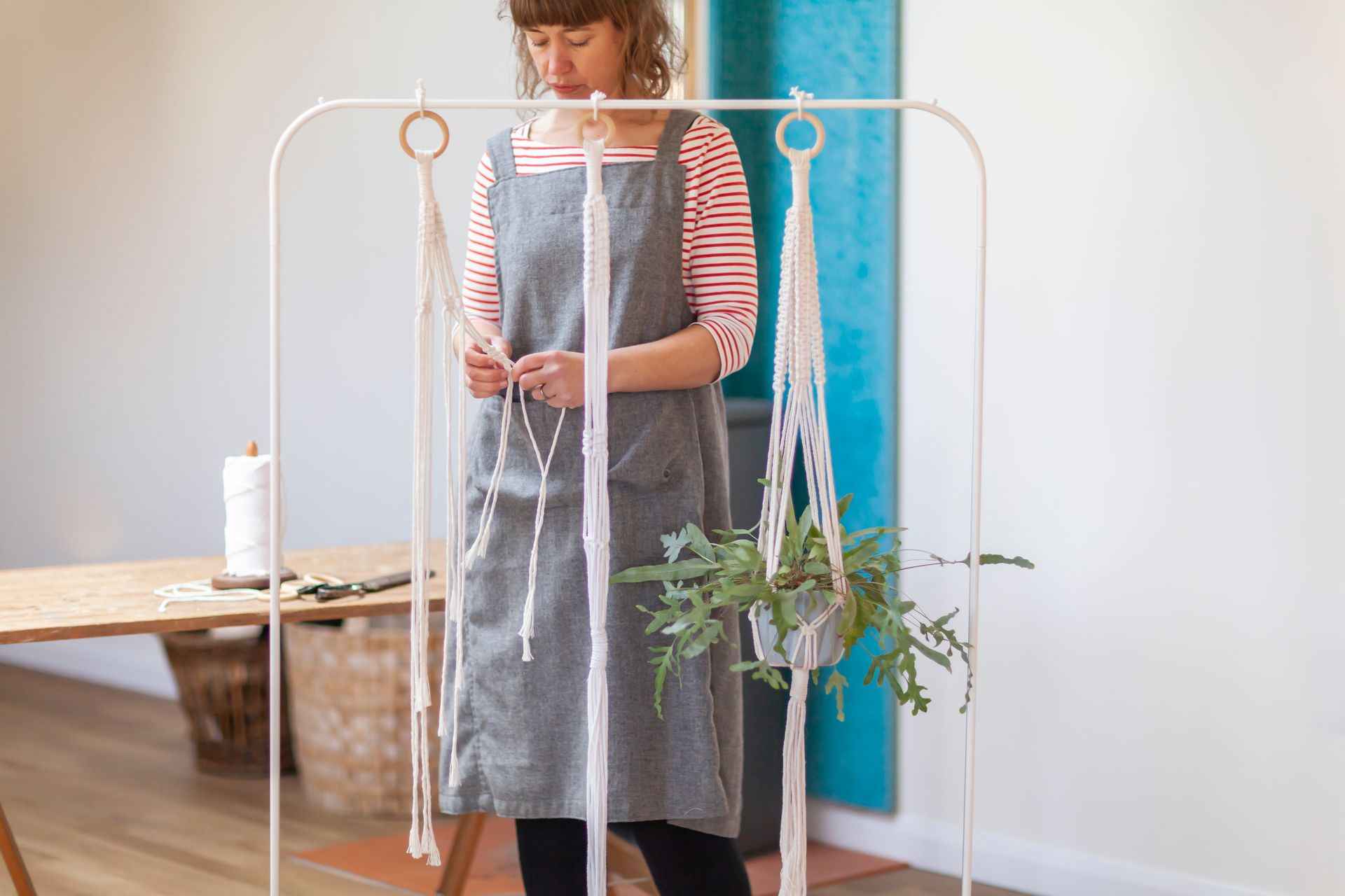 Woman in apron making macrame plant hanger, in light-filled room.
