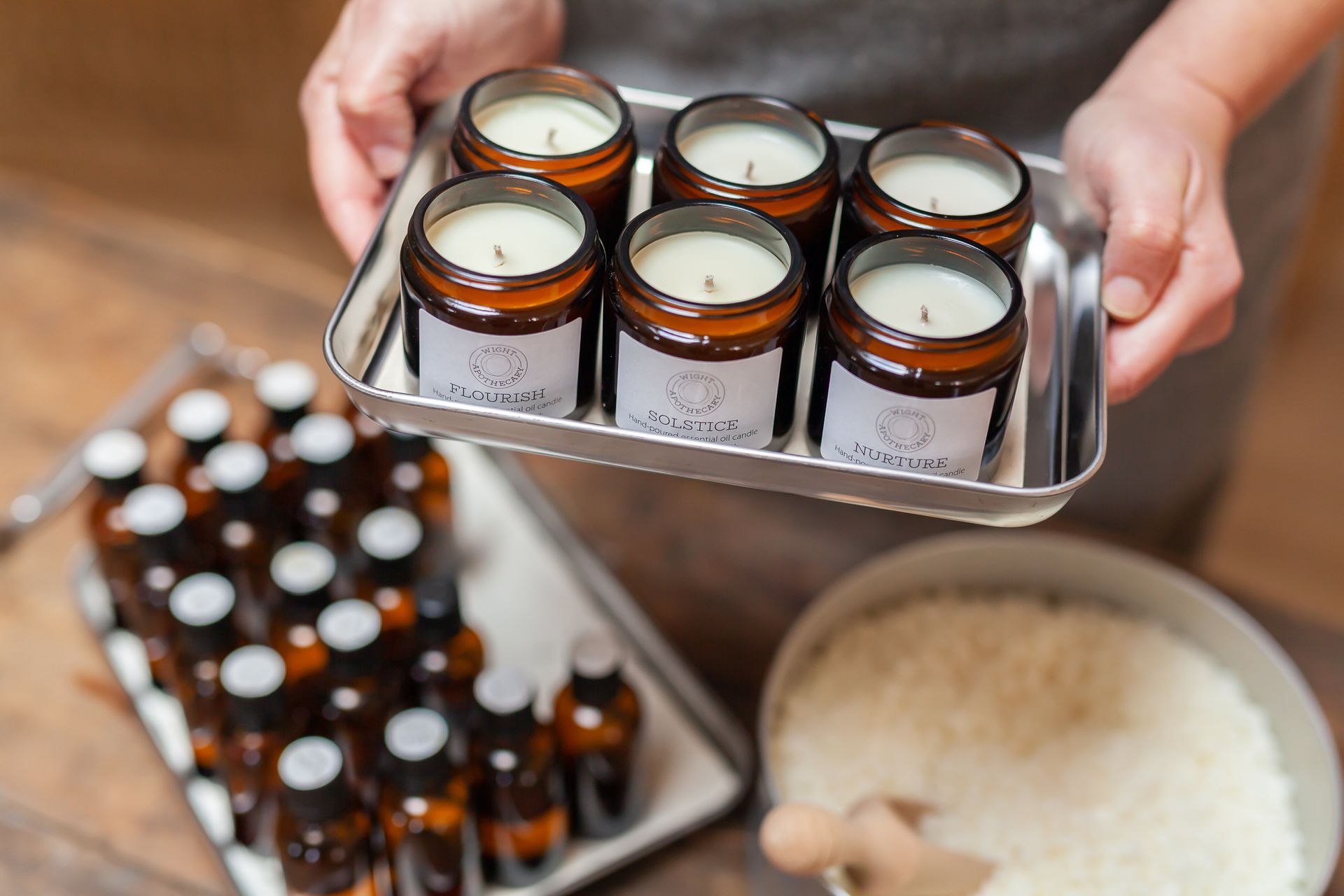 Person holding a tray of six lit candles, with essential oils and wax nearby.