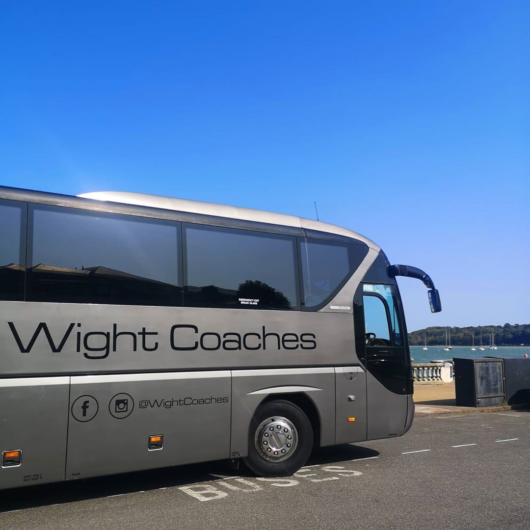 Silver Wight Coaches bus parked near water, under a bright blue sky.